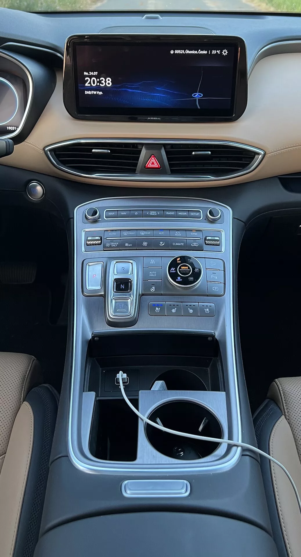 Vista de alto ângulo do interior do console central do Hyundai Santa Fe 2020 a partir dos assentos traseiros. A imagem mostra um painel moderno com uma estrada noturna exibida na tela de infotainment e um relógio digital marcando '20:38.' Abaixo, saídas de ar horizontais elegantes se integram perfeitamente ao painel. A console central apresenta uma variedade de controles táteis e digitais para as funções de clima e multimídia. Um botão rotativo brilhante e interruptores adjacentes sugerem navegação avançada e modos de condução. Mais abaixo, uma área de armazenamento aberta com um cabo branco visível indica conectividade de dispositivos. O túnel central, revestido com acabamento metálico, melhora o design interior luxuoso.