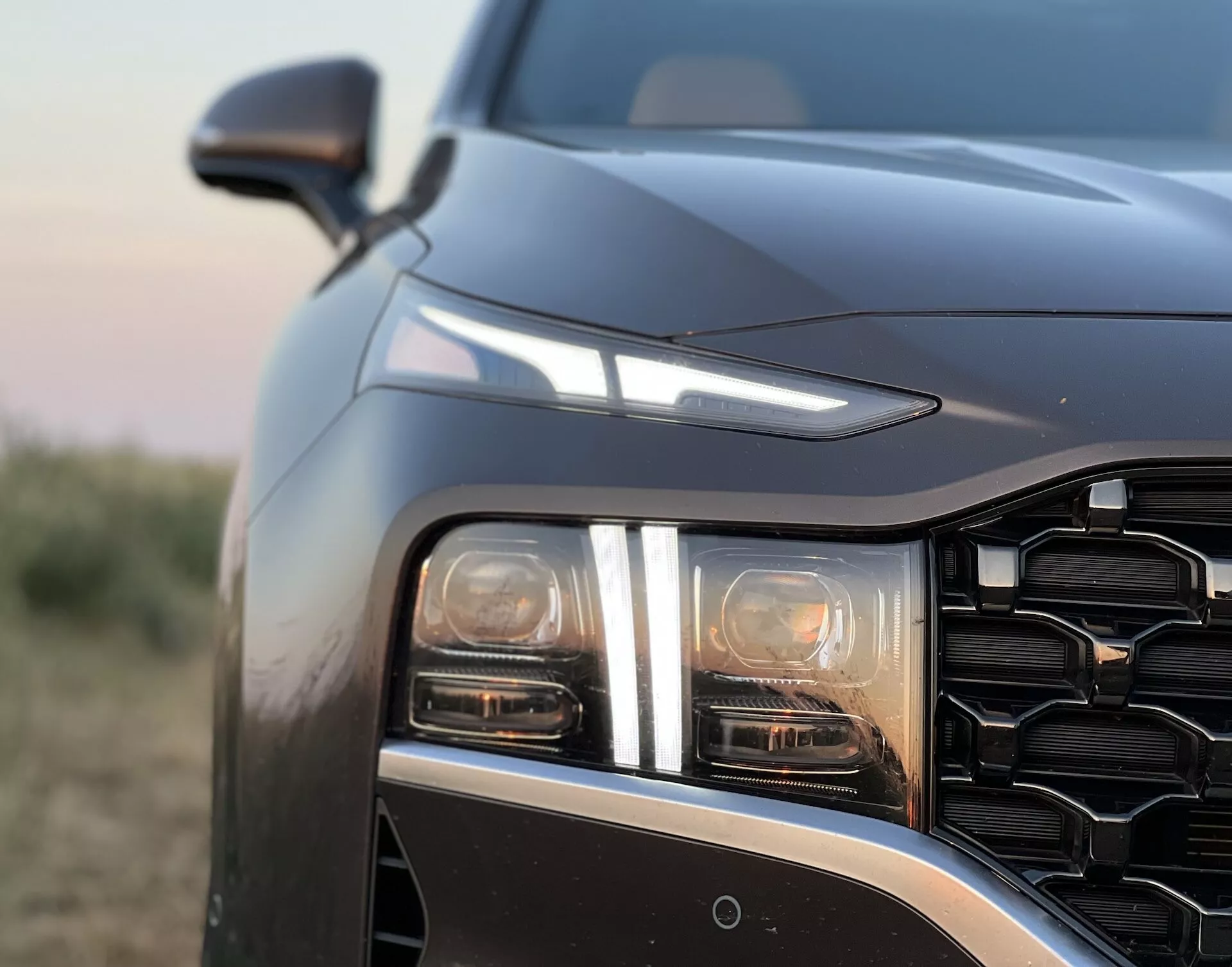 Uma vista detalhada de perto do Hyundai Santa Fe 2020, focando no quarto dianteiro esquerdo a partir de um ângulo ligeiramente frontal e baixo. A imagem enfatiza o conjunto de faróis LED ou HID com pelo menos dois elementos de luz distintos, possivelmente incluindo um farol principal e luz diurna ou indicador de direção. A grade com padrão geométrico e a linha do capô que desce abruptamente sugerem um design contemporâneo e agressivo. O espelho lateral é visível logo além da linha do capô, sugerindo funcionalidades tecnológicas avançadas como um sensor ou câmera no para-choque dianteiro, possivelmente para assistência de estacionamento ou prevenção de colisões. A fotografia, tirada durante o amanhecer ou anoitecer, mostra iluminação de baixo contraste com tons quentes e uma profundidade de campo rasa que destaca os detalhes do design do veículo contra um fundo suavemente desfocado.