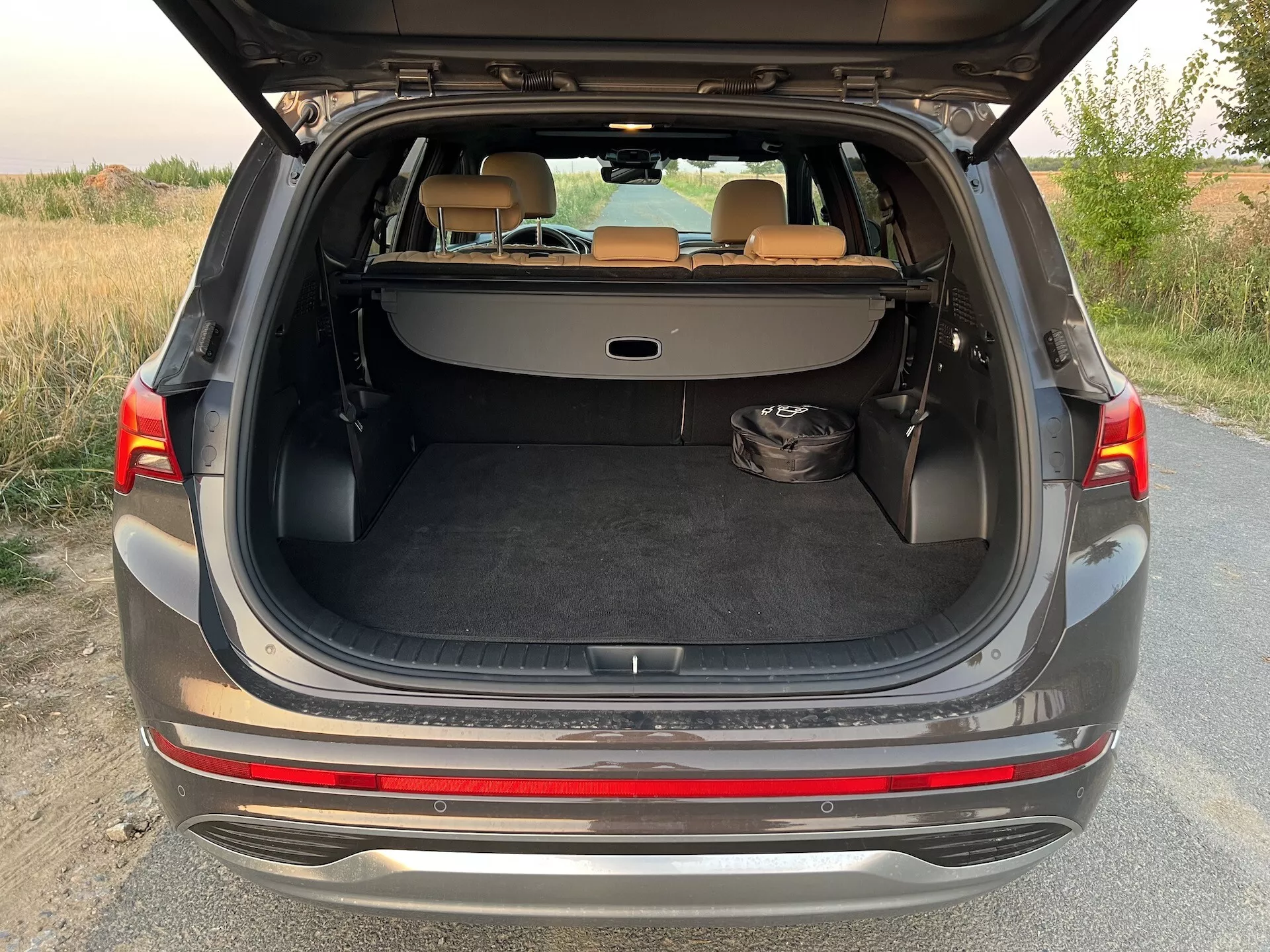 Seção traseira de um Hyundai Santa-Fe 2020 com o porta-malas hatchback totalmente aberto, mostrando o amplo espaço de carga. O veículo, pintado de cinza ou prata, está estacionado em um caminho não pavimentado cercado por vegetação em um ambiente rural ou semi-rural durante a iluminação suave do amanhecer ou anoitecer. O design moderno inclui um para-choque de dois tons, borda de carga cromada protetora, piso de carga plano e pontos de fixação para uma cobertura de carga. Os bancos traseiros estão rebatidos em uma configuração 60/40, proporcionando uma superfície de carga estendida. A área de carga é revestida de carpete e um objeto preto está no lado esquerdo do piso. O interior está em boas condições e as lanternas traseiras estão parcialmente visíveis.