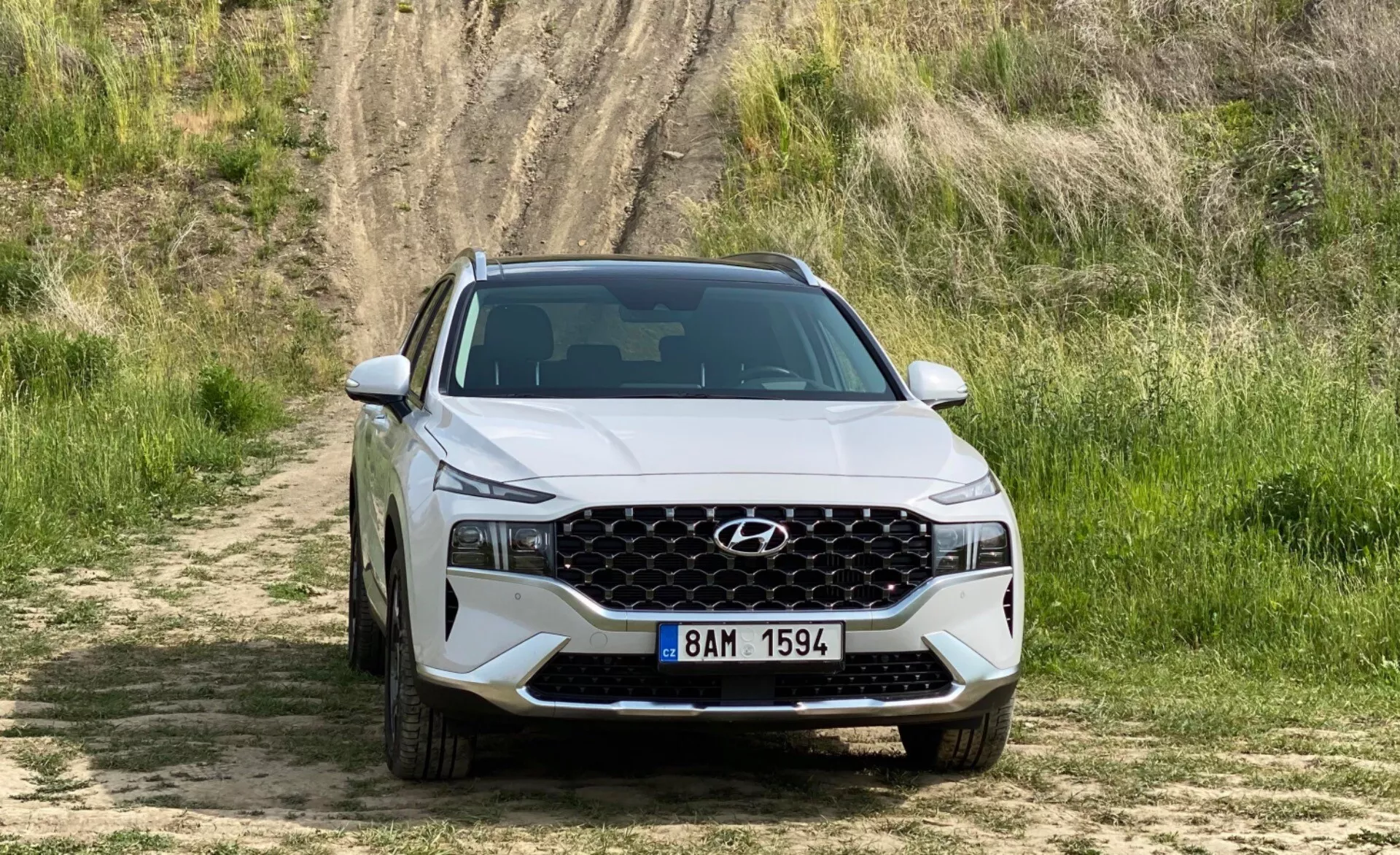 Vista frontal de um Hyundai Santa-Fe 2020 SUV posicionado em um caminho de terra com grama e uma colina ao fundo, mostrando a grade frontal com um padrão de malha geométrica, faróis modernos e afiados com luzes diurnas de LED, e um para-choque dianteiro com entradas de ar. O capô apresenta contornos acentuados e uma placa de matrícula ao estilo europeu é visível. A imagem enfatiza o design e os recursos do veículo em um cenário off-road.