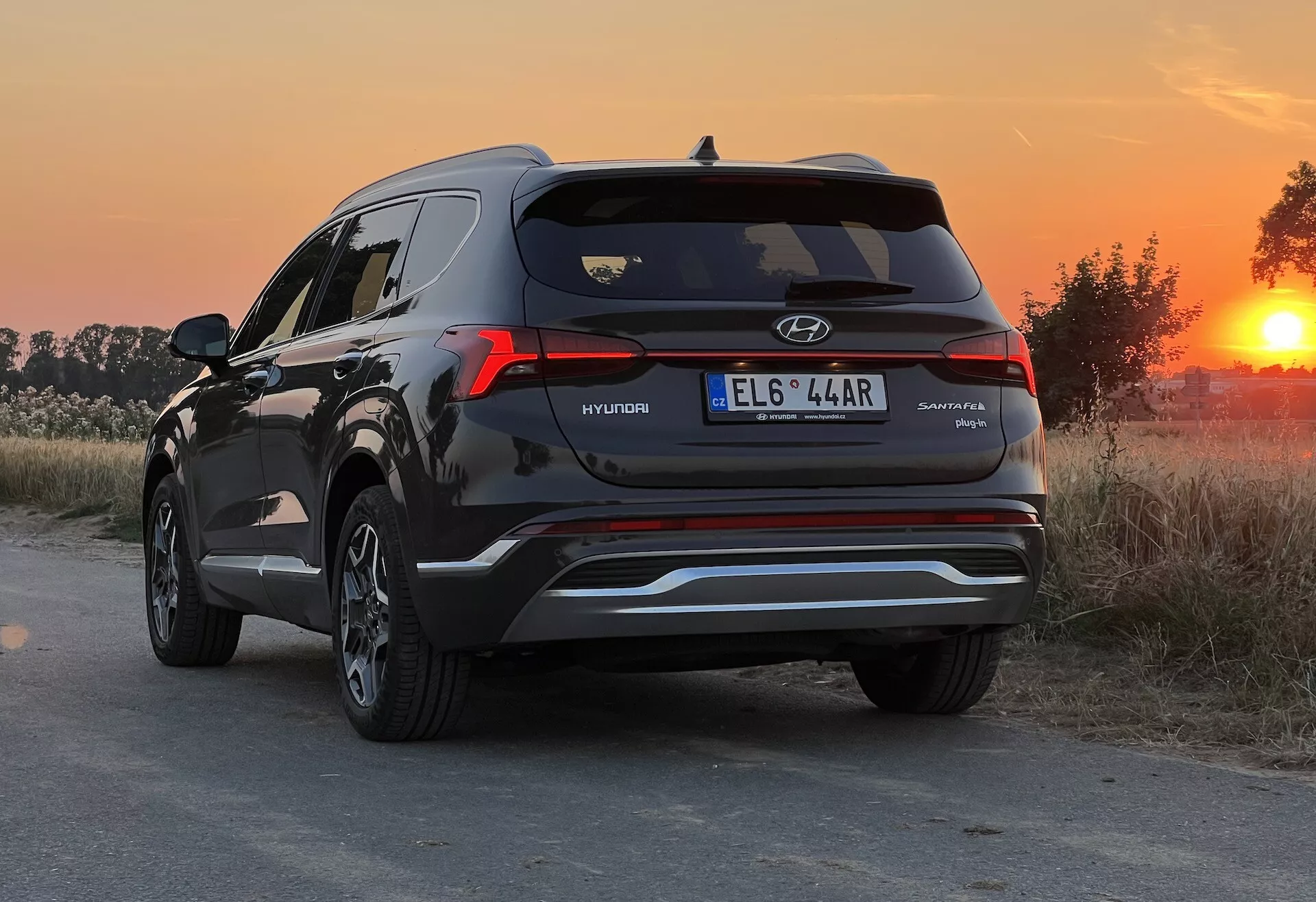 Vista traseira três quartos de um SUV Hyundai Santa Fe 2020, destacando o lado direito e perfil traseiro com insígnia visível. A fotografia foi tirada durante a hora dourada, oferecendo uma luz de fundo quente que acentua a cor da pintura e os reflexos. O SUV está estacionado em um caminho de terra ou não pavimentado com ambiente rural. O ângulo baixo da câmera, em torno da altura das luzes traseiras, mostra o design das luzes traseiras, janela traseira, contornos da tampa do porta-malas e pontas de escape duplas. Também são visíveis detalhes como uma linha metálica no para-choque inferior, um spoiler traseiro e uma antena shark-fin.
