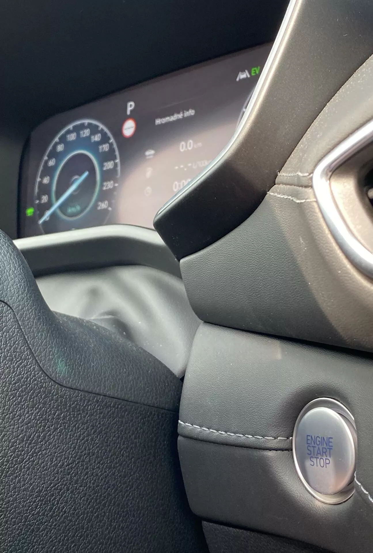 Vista interior em close-up do Hyundai Santa Fe 2020, focando no lado esquerdo do volante revestido em couro e no painel de instrumentos digital. A foto, tirada do banco do motorista e ligeiramente inclinada para cima, destaca o design moderno do volante, costuras detalhadas, pegada contornada e a ignição sem chave com um botão 'ENGINE START STOP'. O painel de instrumentos digital exibe o velocímetro, indicando que o carro está parado, e outras informações do veículo, enfatizando um design premium e ergonômico.