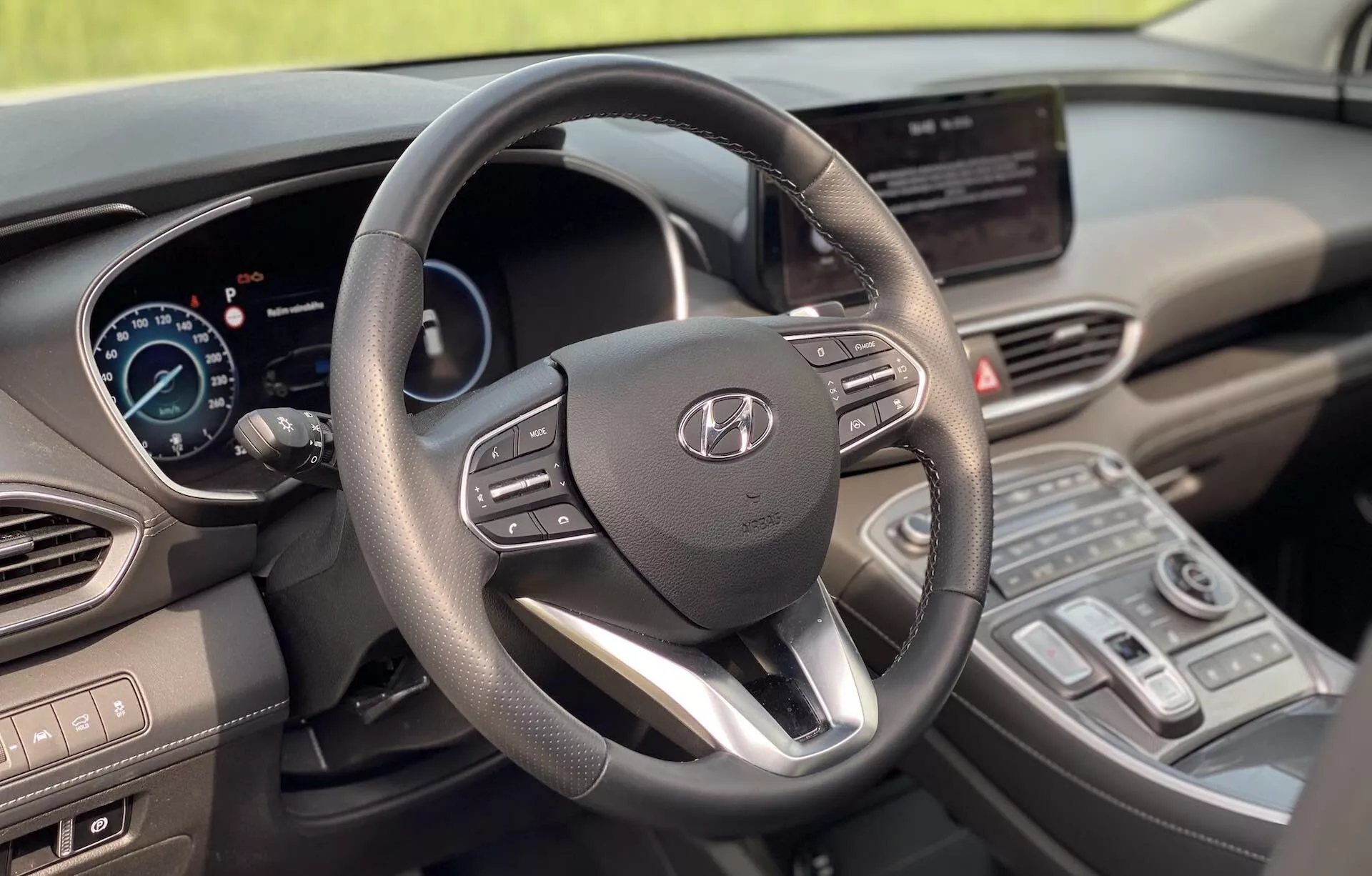 Imagem do interior do Hyundai Santa Fe 2020 a partir do assento do motorista, destacando o volante moderno de três raios com logotipo Hyundai e controles integrados, painel de instrumentos digital, tela central de infotainment touch, consola central com controles de climatização e uma alavanca de câmbio moderna. O design apresenta um equilíbrio entre funcionalidade e estética moderna com materiais suaves ao toque e acentos metálicos, indicando características tecnológicas avançadas. A imagem mostra um veículo limpo, bem conservado e estacionário, sem desfoque de movimento.