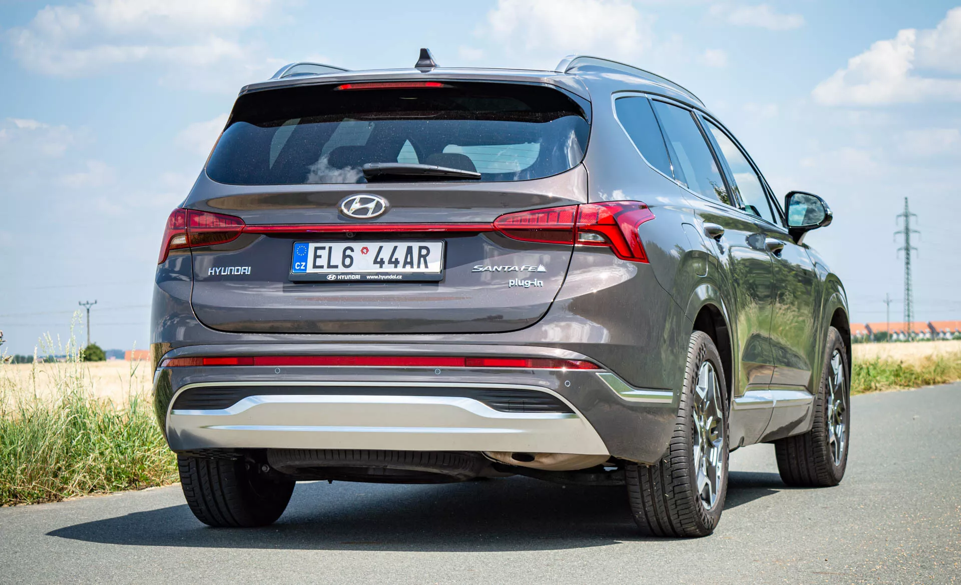 Vista de ângulo traseiro do SUV Hyundai Santa Fe Hybrid 2020 com emblema Hyundai no porta-malas, marca SANTA FE, luzes traseiras LED distintivas, spoiler traseiro, placa de proteção prateada, placa de licença central, rodas de liga leve e acabamento de pintura metálica, capturada em um dia claro com uma estrada asfaltada e áreas gramadas.