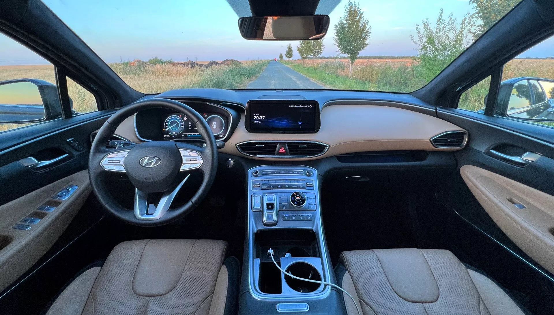 Vista de grande ângulo do assento do condutor de um Hyundai Santa Fe 2020 que mostra os bancos de couro bege, o painel de instrumentos detalhado com cluster digital, volante com marca Hyundai e controles montados, sistema de infoentretenimento com tela e uma consola central totalmente equipada com alavanca de câmbio, porta-copos, botões de controle e compartimento de armazenamento.