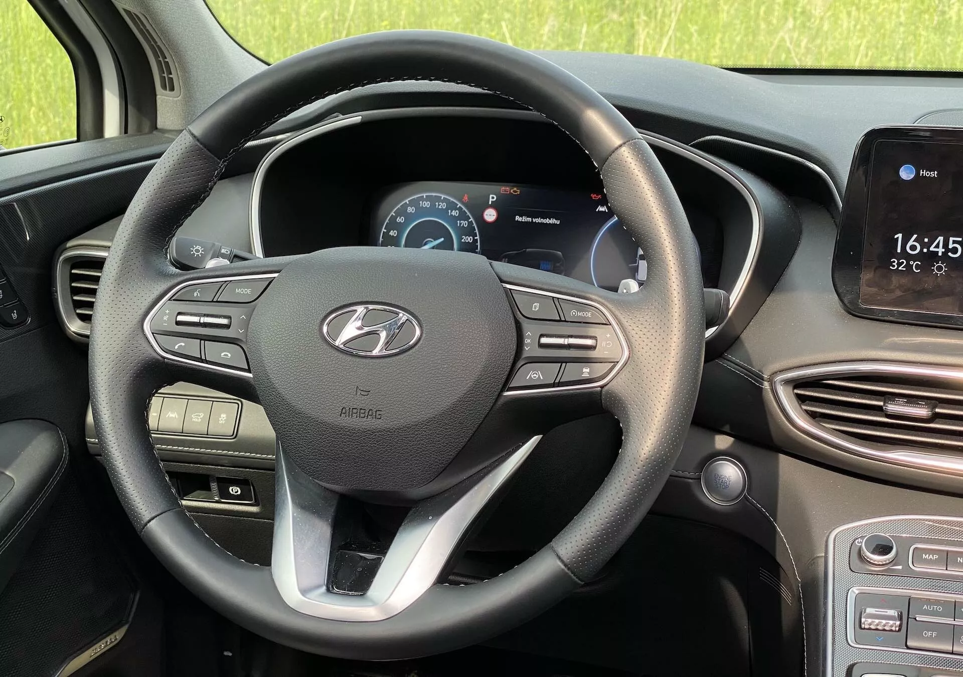 Vista interior de um Hyundai Santa Fe 2020 a partir da perspectiva do banco do condutor, mostrando o volante moderno de três raios com acento prateado e logotipo Hyundai. O raio esquerdo possui controles de volume e mídia, enquanto o raio direito apresenta botões de controle de cruzeiro e navegação de menu. Atrás do volante, um painel de instrumentos digital exibe a velocidade e o tacômetro. À direita está uma tela sensível ao toque do sistema de infoentretenimento exibindo hora e temperatura, com navegação potencial e integração de smartphone. Abaixo estão aberturas de ventilação e botões de controle do clima. O carro está estacionado à luz do dia, com um ambiente natural e não urbano visível através das janelas.