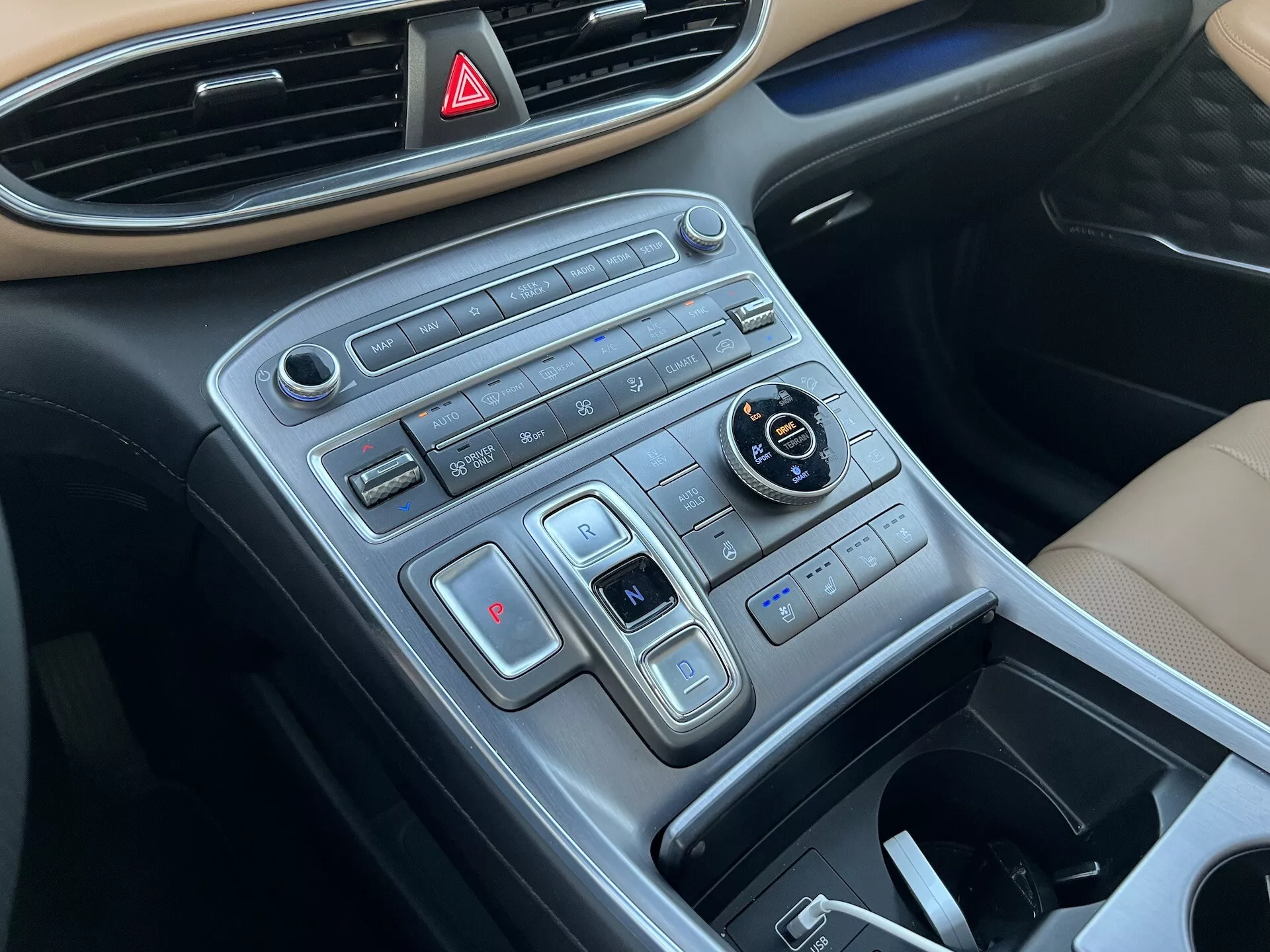 Visão aproximada da consola central do Hyundai Santa Fe 2020 a partir do ângulo do lado do passageiro, mostrando o painel de instrumentos com o botão das luzes de emergência na parte superior, controlos de design moderno em filas horizontais que incluem o seletor de modo de condução, configurações do controlo de tração, sistema de climatização de dupla zona, botões de aquecimento dos bancos e um compartimento de armazenamento retrátil, com acabamento que imita metal escovado.