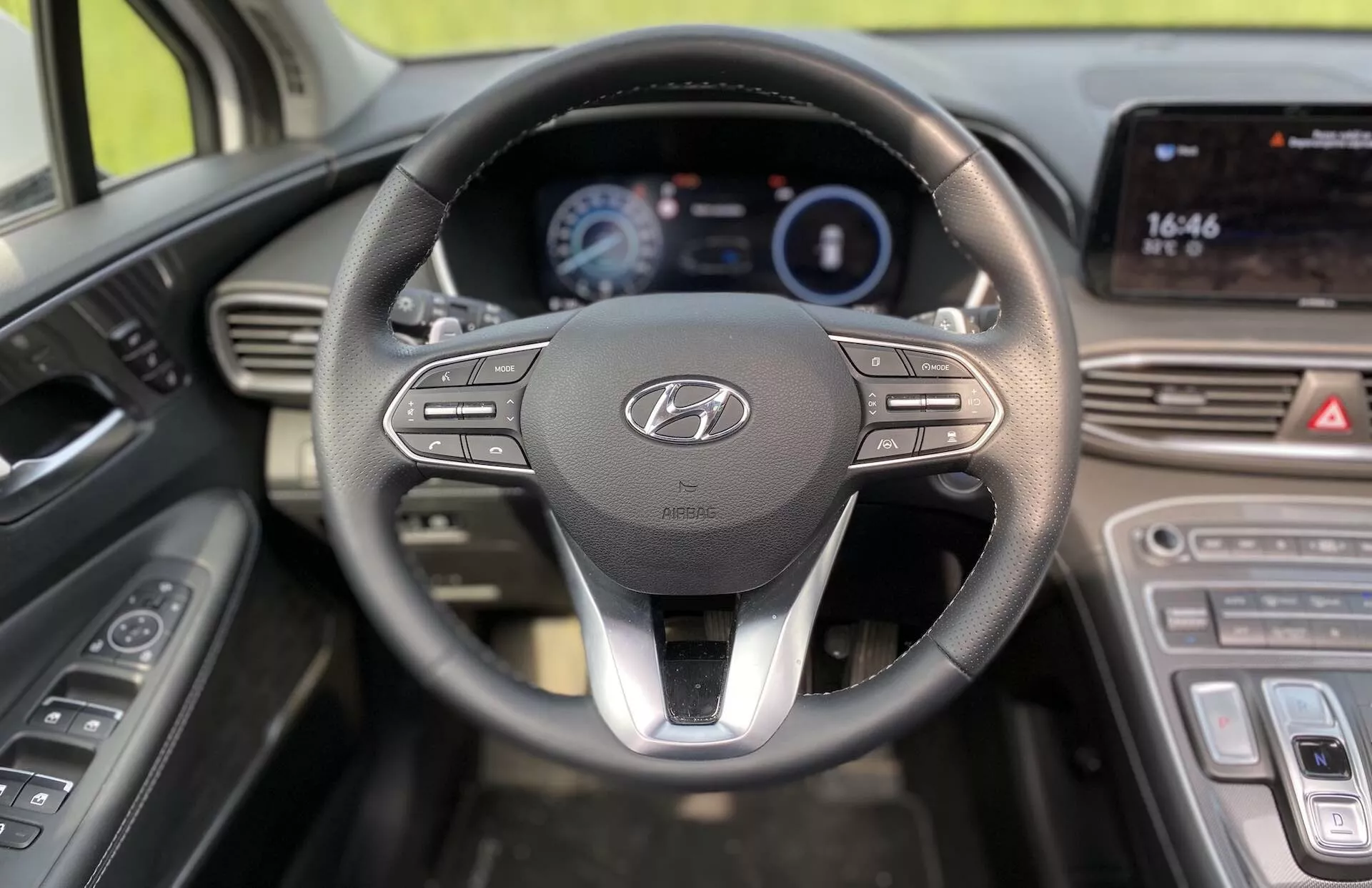 Perspectiva do motorista do interior do Hyundai Santa-Fe 2020 modelo híbrido, com um volante proeminente apresentando o logotipo da Hyundai e botões de controle para multimídia e conectividade. Atrás do volante há um painel de instrumentos digital iluminado que exibe informações importantes, como velocidade e economia de combustível. À direita, o console central inclui um display touch screen exibindo a hora (16:46) e provavelmente suporta funcionalidades de navegação e multimídia. Abaixo do display touch screen, controles de clima sensíveis ao toque são visíveis, melhorando o design moderno do veículo. Controles de porta para vidros elétricos e ajustes dos espelhos laterais podem ser vistos à esquerda do volante.