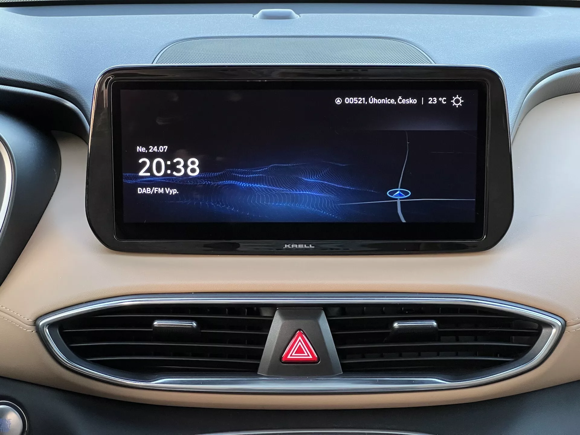 Close-up da área central do painel de um Hyundai Santa Fe 2020, destacando a tela horizontal e ampla do sistema de infotainment, mostrando um relógio digital '20:38', fonte de mídia rotulada como 'DAF/MP3', leitura de temperatura '23°C' e texto parcialmente visível em polonês. Também são visíveis duas saídas de ar com detalhes cromados e o botão de luzes de emergência abaixo da tela. O painel apresenta um esquema de cores bicolor com uma estética interior moderna e de alta qualidade.