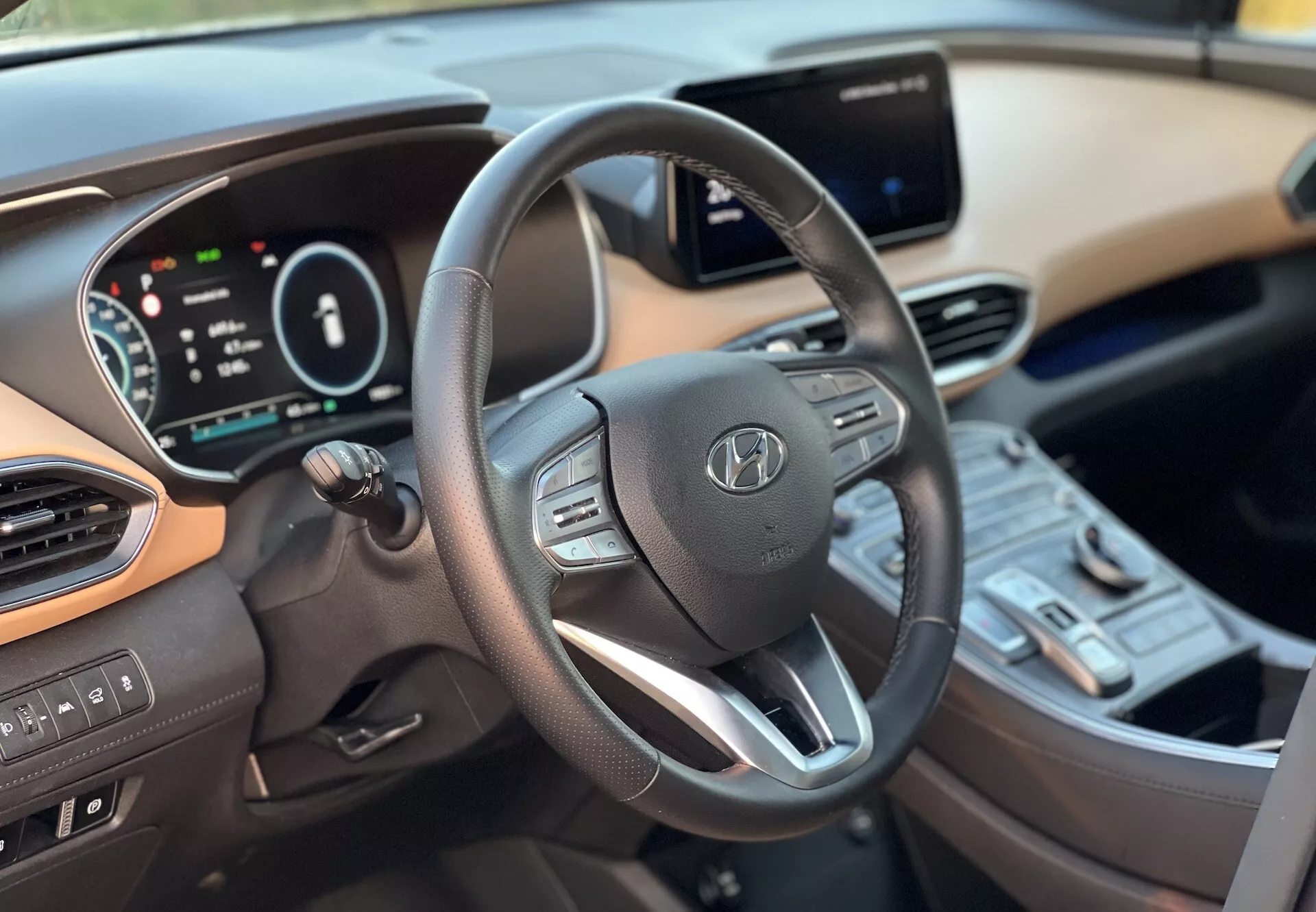 Visão interna do Hyundai Santa-Fe 2020 mostrando o lado do motorista de um ângulo ligeiramente à direita do centro, focando no volante moderno de três raios com controles embutidos, painel de instrumentos digital e um sistema de infotainment touchscreen no console central. Interruptores de controle climático, botão de partida e esquema de cores dual do painel e interiores também são visíveis, ilustrando a tecnologia avançada e o conforto do veículo.