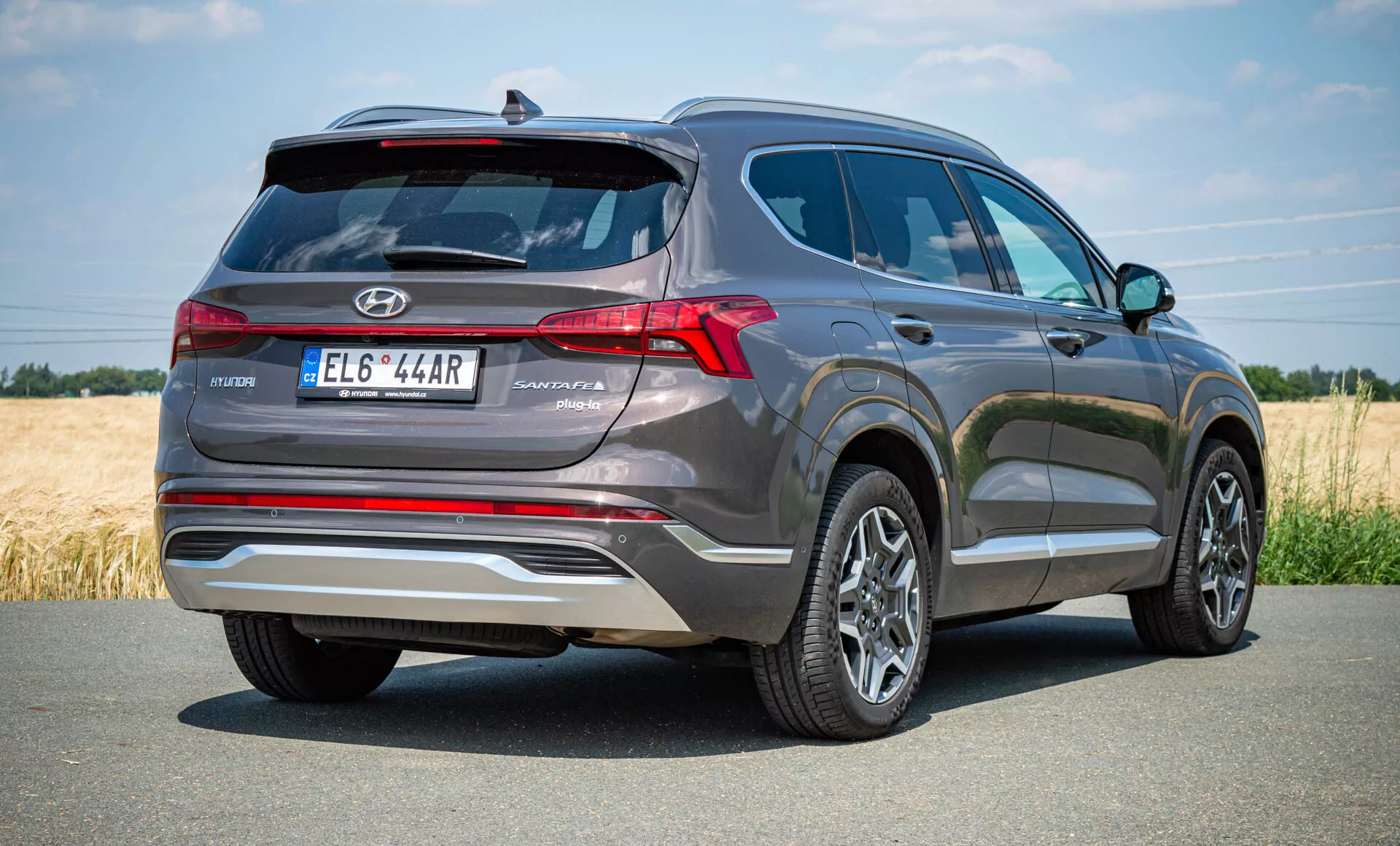 Visão traseira de três quartos de um Hyundai Santa Fe Hybrid 2020. A foto em ângulo baixo enfatiza a altura e a robustez do SUV, com o carro posicionado no lado direito sugerindo movimento. Estacionado em uma estrada pavimentada em um ambiente rural, a traseira apresenta lanternas traseiras de LED dispostas verticalmente, um spoiler traseiro para estabilidade aerodinâmica, pontas de escape duplas, spoiler de teto dividido e acabamentos refletivos no para-choque. O design sugere a presença de sensores de estacionamento traseiros, câmera de ré, rodas de liga leve multifachadas e uma antena barbatana de tubarão para rádio via satélite ou tecnologia GPS.