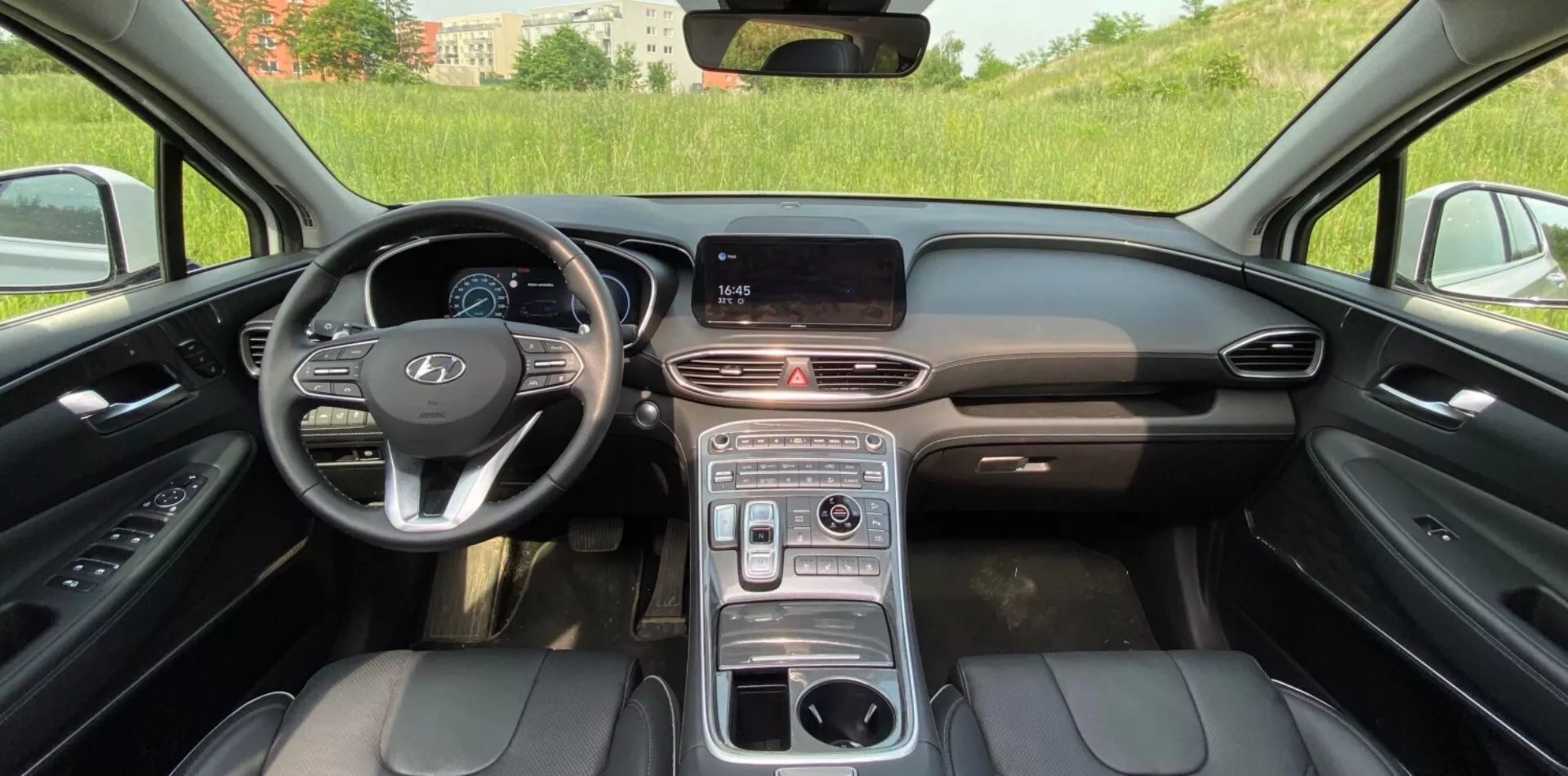 Vista do interior do Hyundai Santa Fe 2020, destacando a cabine dianteira moderna com um painel central com display digital de infotainment, tira de ventilação elegante e botões multifuncionais no volante. O design do volante à esquerda inclui o logo proeminente do fabricante e provavelmente um painel de instrumentos digital. A consola central possui um seletor de marchas rotativo, suportes para copos e possivelmente um apoio de braço. Os assentos dianteiros são estofados em couro preto com costuras visíveis, sugerindo ajustes elétricos e luxo. A configuração parece ser ao ar livre com luz natural do dia, com um campo verde ligeiramente desfocado visível através do para-brisa.
