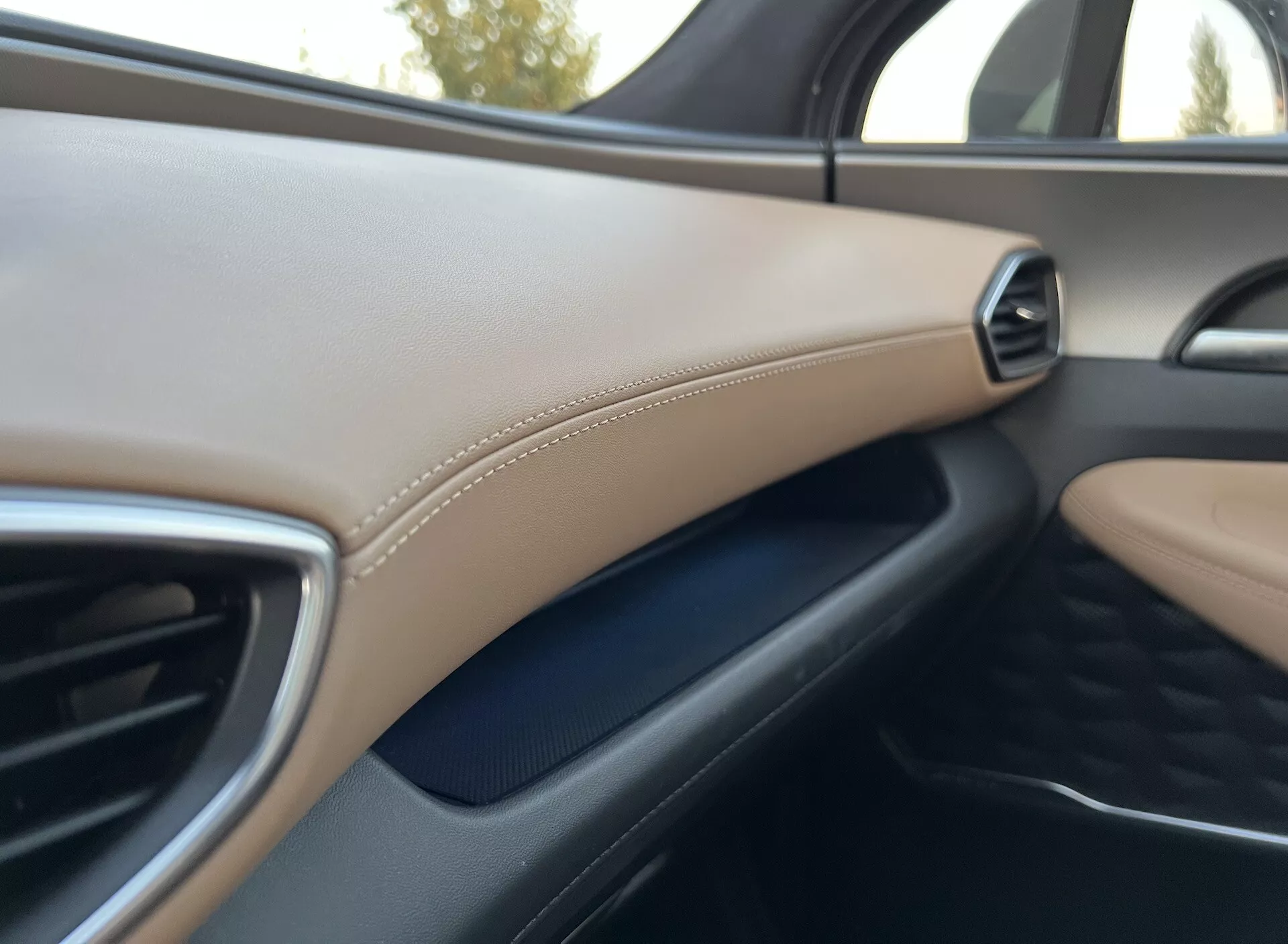 Detalhe do interior do Hyundai Santa Fe 2020 mostrando o lado do painel do passageiro. A imagem, tirada do banco do motorista ou do passageiro central, destaca uma ventilação lateral direita com acabamento metálico. Abaixo da ventilação, é visível uma superfície texturizada cinza escuro, enquanto um painel bege com costura decorativa acentua o acabamento de luxo. O foco é suave, dando uma impressão geral do design interior, materiais e qualidade de construção. A luz do dia pode ser inferida pela janela, embora os detalhes exteriores não sejam claros.