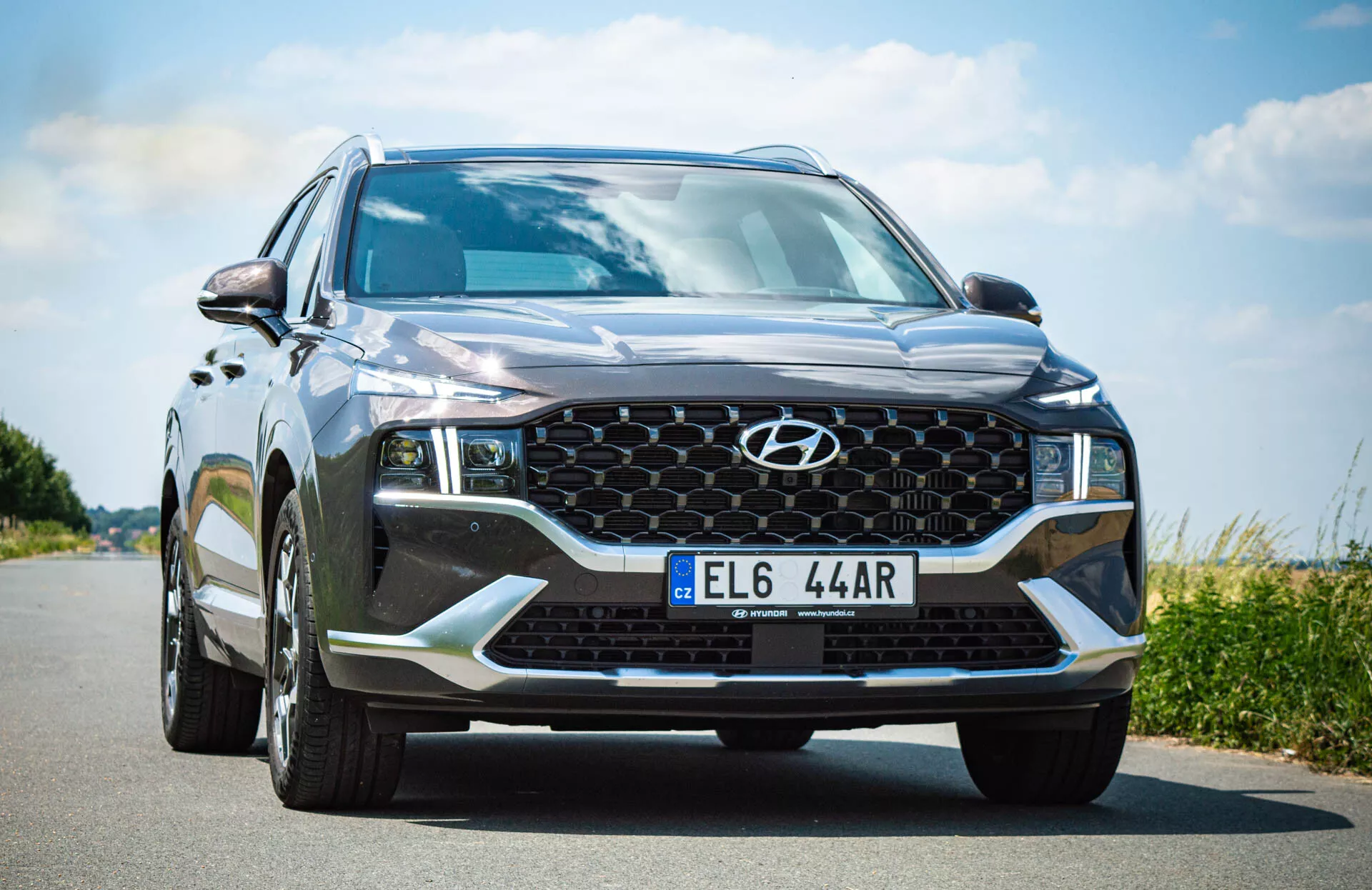 Vista angular frontal de um SUV moderno, especificamente um Hyundai Santa Fe 2020, tirada de um ponto baixo próximo ao lado do motorista. A câmera captura o ousado design frontal do veículo, incluindo uma cor metálica preta ou cinza escuro com um brilho lustroso, uma grade em padrão de favo de mel com um emblema Hyundai central, faróis angulares e afiados com luzes diurnas de LED, iluminação adicional no para-choque inferior, como faróis de nevoeiro, uma grande entrada de ar e linhas esculpidas no capô. O para-brisa é grande e inclinado, com espelhos laterais parcialmente visíveis e sinais de direção integrados. A placa de licença está montada centralmente na parte inferior do para-choque.