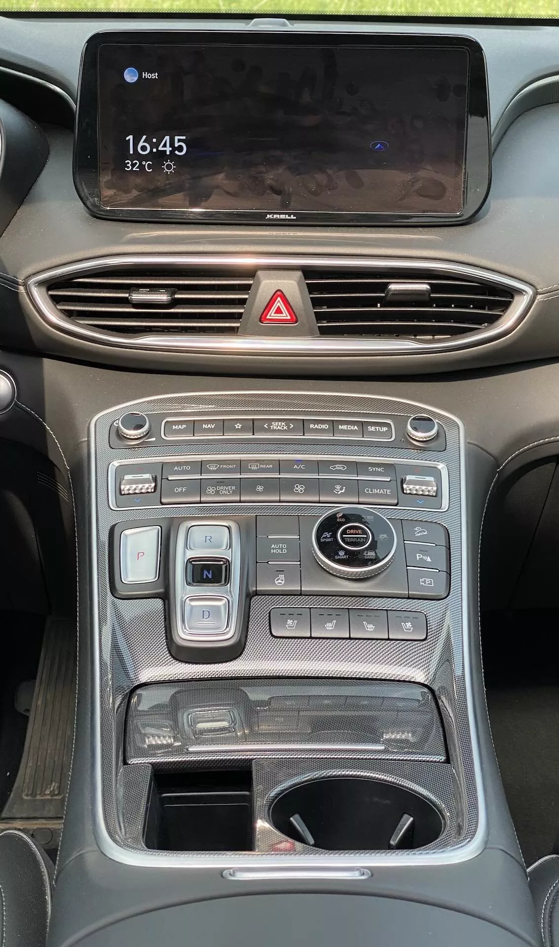 Imagem do console central do interior de um Hyundai Santa Fe 2020, destacando diversos controles e telas no painel a partir de uma perspectiva ao nível dos olhos. Na parte superior, há uma tela sensível ao toque de infotainment em orientação horizontal exibindo '16:45' e '32°C', com uma superfície brilhante que reflete o interior. Abaixo estão as saídas de ar horizontais com detalhes cromados e um botão de luzes de emergência vermelho. O console inclui botões e dials, incluindo um sistema de controle climático de dupla zona, controles de temperatura dos bancos e mais controles de funções do veículo, como modos de condução e sensores de estacionamento. As alças em ambos os lados enfatizam esportividade ou capacidade off-road.