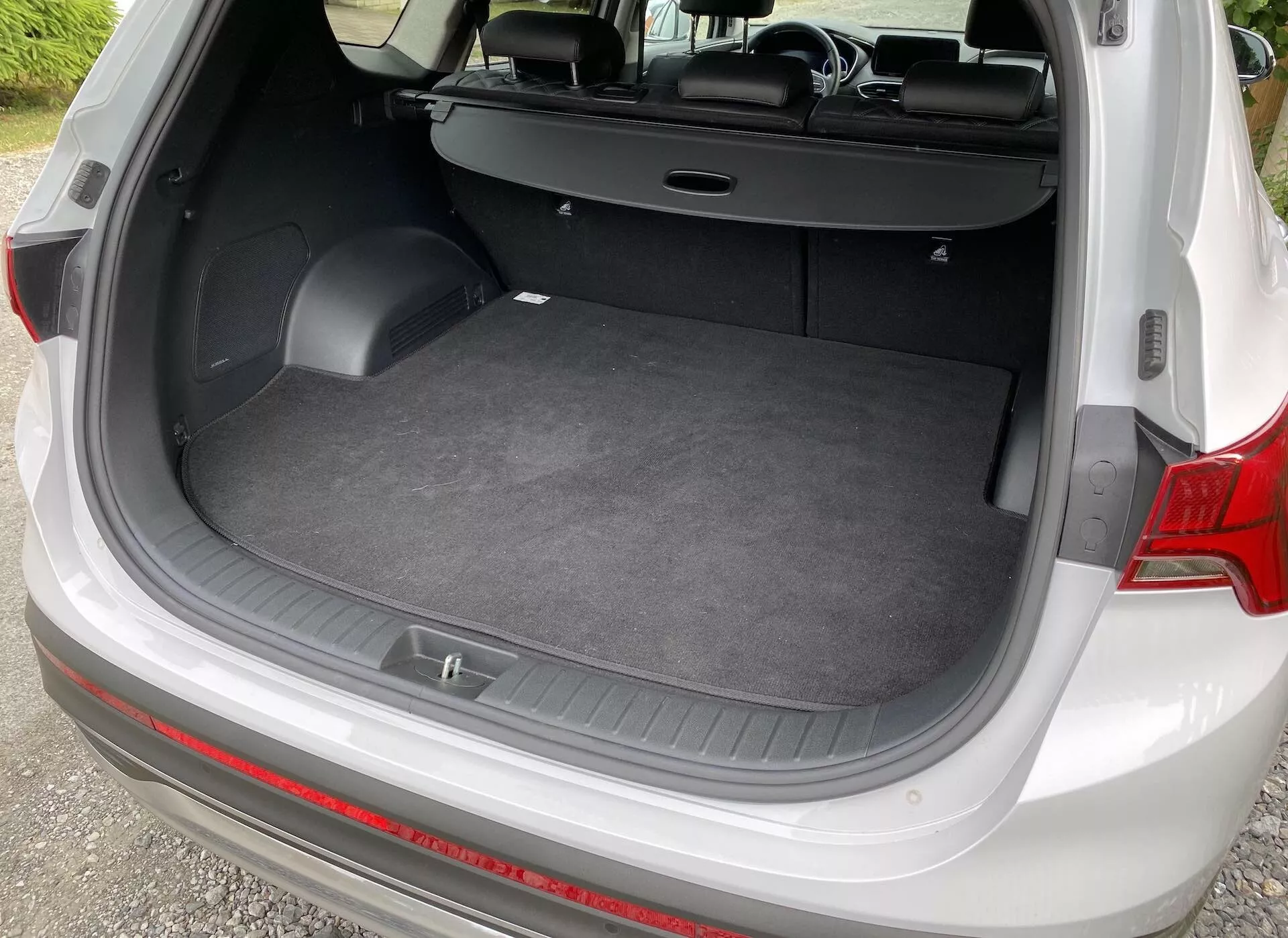 Foto em ângulo alto da área de carga de um Hyundai Santa-Fe 2020, vista de trás com a porta traseira aberta. A imagem mostra um piso de carga plano, superfície acarpetada e paredes laterais com pequenos compartimentos de armazenagem e ganchos para acessórios. Os bancos traseiros estão dobrados na vertical e a capa retrátil do porta-malas está empurrada para trás, destacando o interior versátil do veículo.