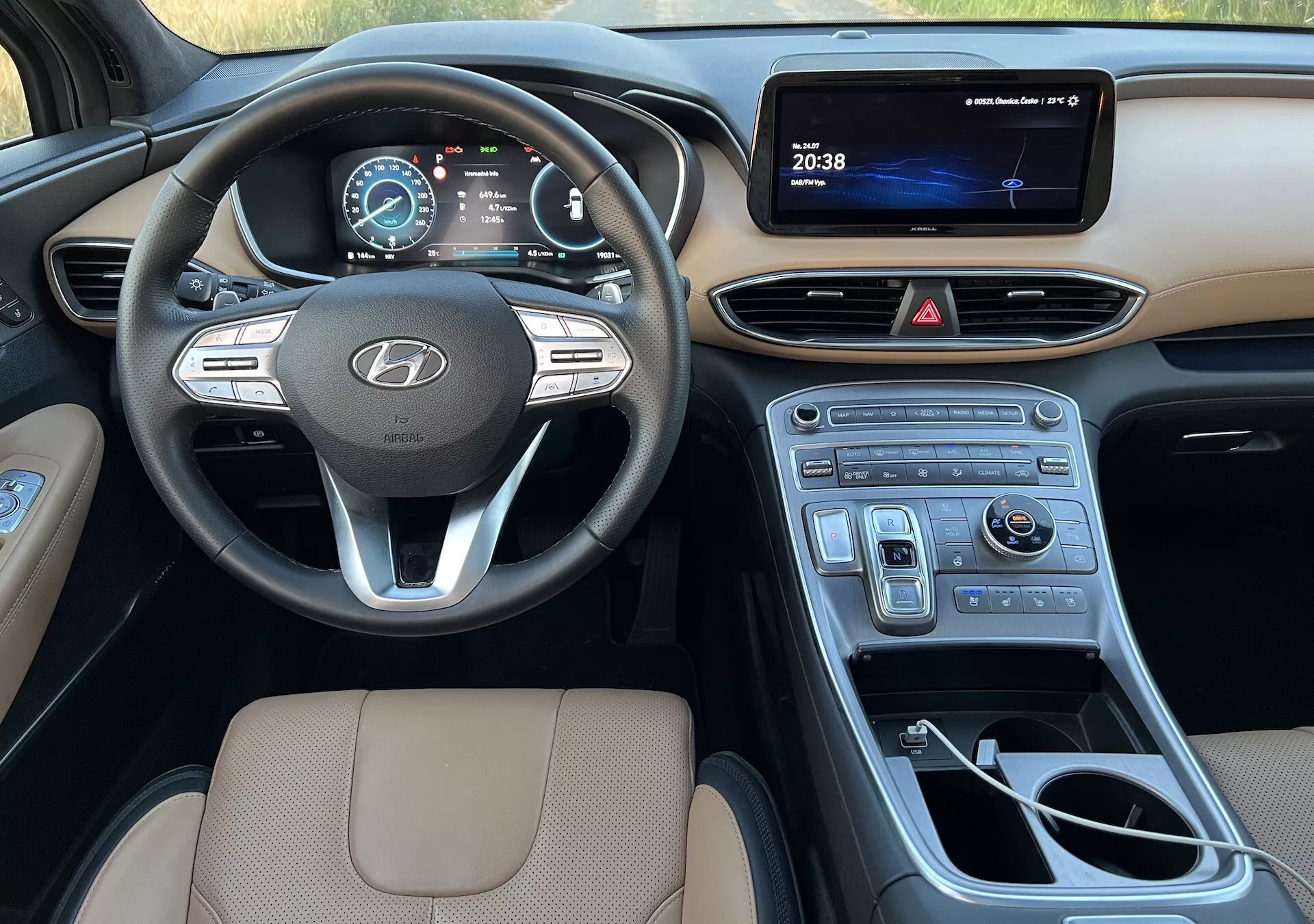 Esta imagem mostra o interior de um Hyundai Santa-Fe 2020, focando-se no lado do condutor. Oferece uma visão abrangente desde o assento do condutor até ao console central, estendendo-se para o lado do passageiro, destacando vários elementos-chave. O volante com o logotipo da Hyundai, com controles para áudio, controle de cruzeiro e sistema de telefone mãos-livres, é mostrado de forma proeminente. Atrás dele, um painel de instrumentos digital exibe o velocímetro e informações do veículo. O console central inclui uma tela de infotainment, botões de controle para luzes de emergência e sistema de controle de clima, e um acabamento brilhante semelhante a madeira. O manípulo da caixa de marchas automática, um botão de controle para o sistema de navegação ou multimídia e porta-copos também estão visíveis. A parte superior do assento do condutor, estofada em material bege com costuras, enfatiza o conforto e a qualidade do design. A imagem captura o design moderno, amigável para o utilizador e as características tecnológicas do interior do veículo durante o dia com iluminação natural.