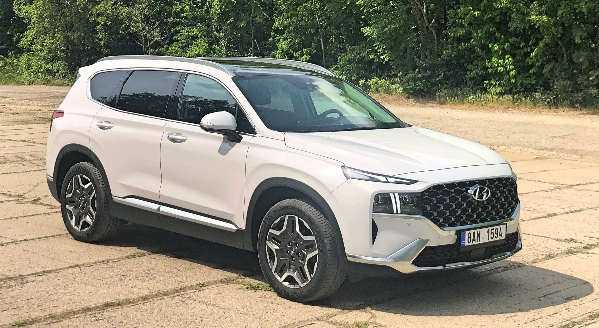 SUV moderno Hyundai Santa Fe 2020 estacionado em superfície de asfalto visto de três quartos frontais, mostrando design de grade, faróis elegantes, rodas de liga leve multirraios, espelhos laterais na cor da carroceria com indicadores de direção integrados, linhas de caráter, perfil aerodinâmico, pintura branca brilhante, vidros ligeiramente escurecidos e iluminação natural e uniforme com vegetação ao fundo.