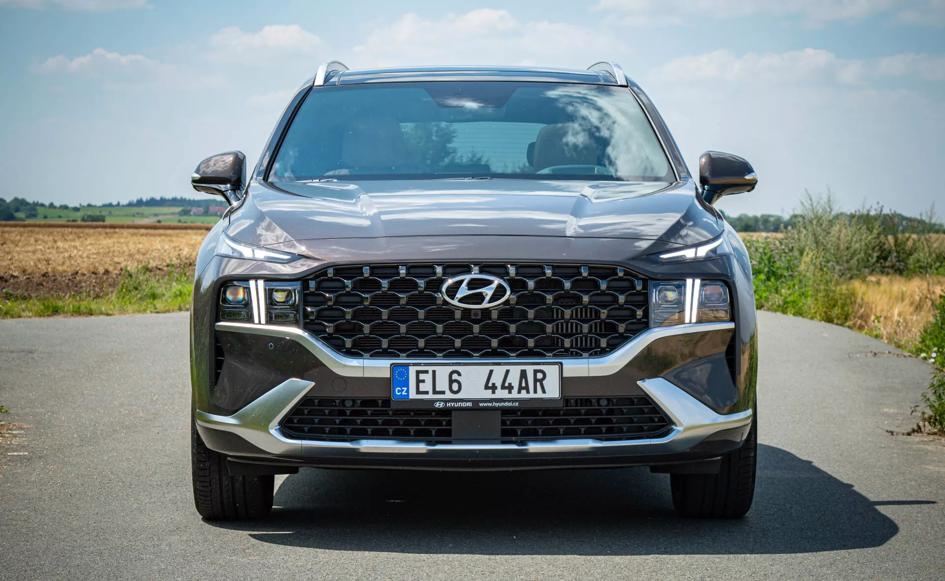 Hyundai Santa Fe 2020 - Vista frontal de um SUV moderno em uma estrada de asfalto com uma paisagem natural ao fundo. Esta imagem em ângulo baixo enfatiza a postura imponente do SUV, destacando detalhes como a grade frontal hexagonal com padrão em favo de mel, faróis de LED angulares, acentos contrastantes no para-choque dianteiro e elementos de iluminação adicionais, incluindo faróis de neblina.