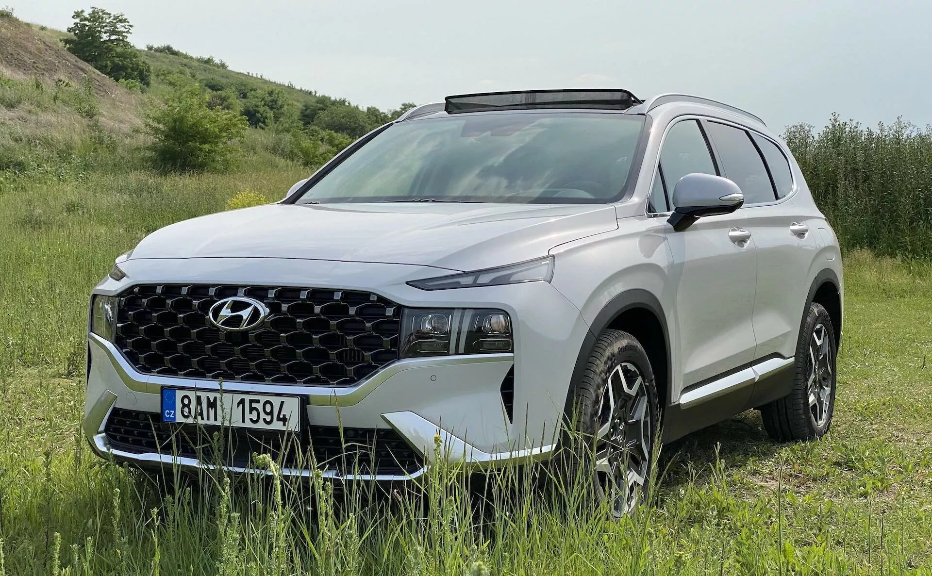 Um SUV Hyundai Santa Fe 2020 moderno em um ambiente natural ao ar livre. O veículo está estacionado em uma área de grama com uma leve inclinação, sugerindo capacidades off-road. Foto diurna brilhante mostrando o perfil dianteiro e lateral com características como uma grade geométrica proeminente, faróis LED estreitos, luzes diurnas, rodas de liga leve com design de múltiplos raios, espelhos laterais na cor da carroceria com sinais de seta integrados, trilhos de teto e revestimento plástico ao redor dos arcos das rodas. A imagem captura o design e o potencial aventureiro do SUV, destacando sua utilidade e aparência premium.