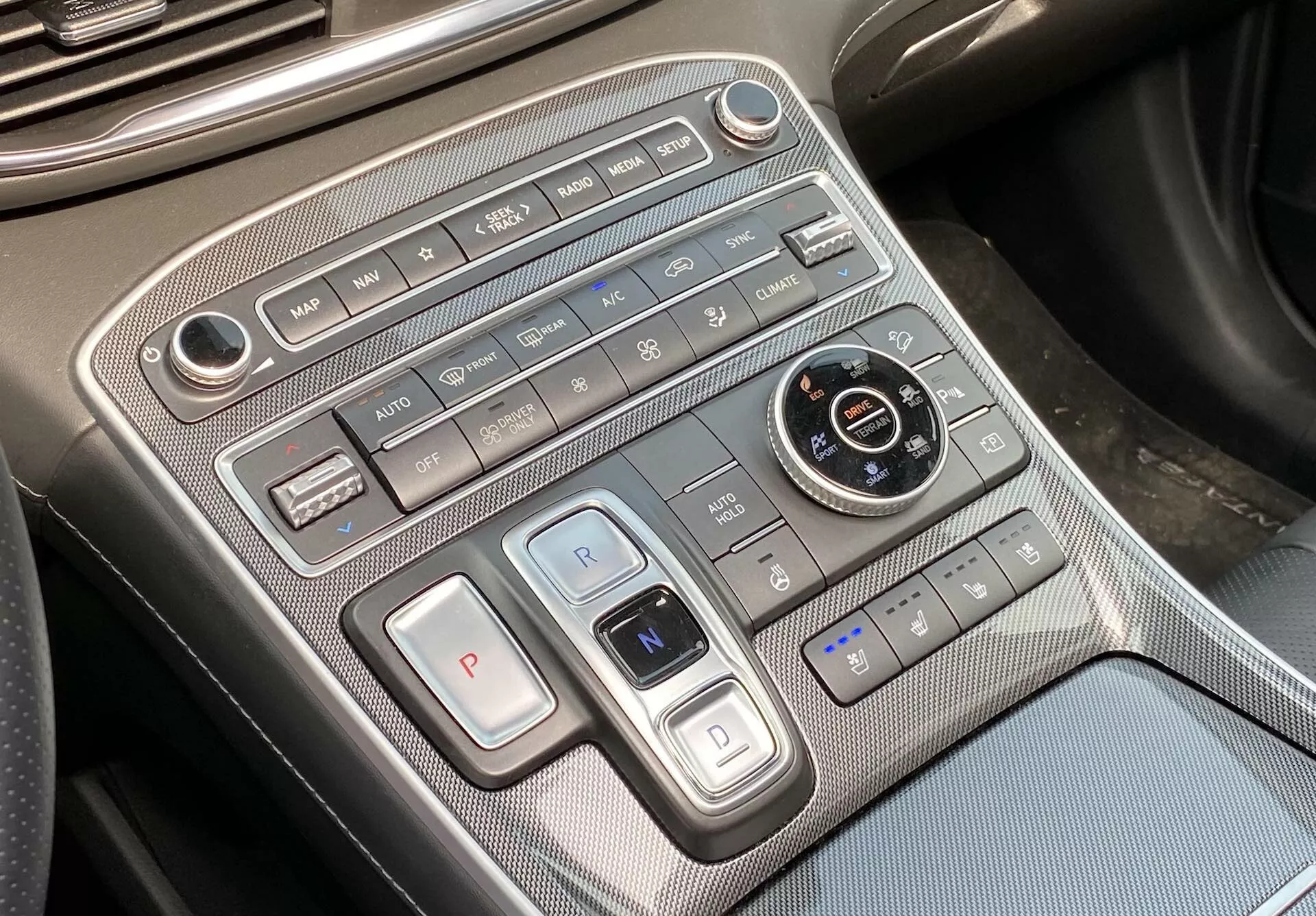 Esta imagem mostra uma visão de close-up do console central do Hyundai Santa Fe 2020, focando na área de controle do clima e da transmissão. A foto, levemente angulada da perspectiva do motorista ou passageiro, revela um painel de controle do clima disposto horizontalmente na parte superior, incluindo controles de velocidade do ventilador, botões de fluxo de ar, controle de clima automático, desembaçamento e recirculação de ar. Abaixo, existem botões de seleção de marchas com um grande botão 'P' para estacionamento e uma alavanca semelhante a um interruptor para as marchas 'R' e 'N', indicando o uso de um sistema de botão ou eletrônico para a seleção de marchas. Há também um dial redondo para seleção de modos de condução, rodeado por botões de manutenção automática do freio, assistente de estacionamento e botões do sistema de partida/parada automática. Os controles dos assentos aquecidos ou ventilados são visíveis no canto inferior direito. O console apresenta materiais premium com acabamentos metálicos e um padrão semelhante à fibra de carbono. Uma porta USB visível no canto inferior esquerdo oferece opções de conectividade para dispositivos.