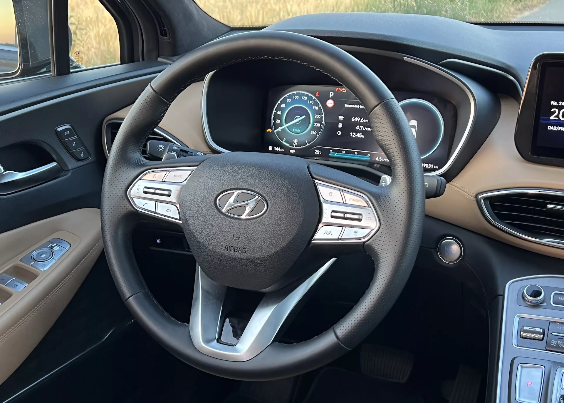 Imagem do interior do Hyundai Santa Fe 2020 focada no volante multifuncional de três raios e no painel de instrumentos digital. Foto tirada da perspectiva do assento do motorista, mostrando os botões de controle de mídia e controle de cruzeiro no volante, o emblema da Hyundai com o texto 'AIRBAG' e um painel digital multifuncional indicando que o veículo está em modo de estacionamento com um aviso de porta aberta. À esquerda estão os controles de luzes e, à direita, é visível a borda de uma tela de infoentretenimento junto às saídas de ar.