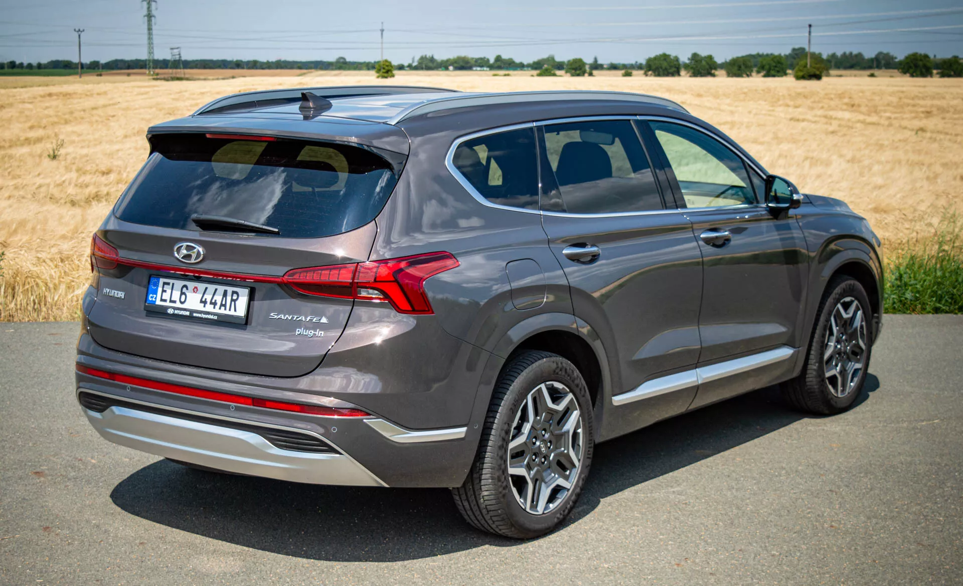 A imagem mostra uma vista traseira de três quartos de um moderno SUV Hyundai Santa Fe 2020, capturada à luz do dia sob céu limpo. O carro está ligeiramente inclinado para a direita, revelando características importantes como o portão traseiro com logotipo da marca, nome do modelo e nível de acabamento, luzes traseiras de LED de design nítido, rodas de liga com design multirraio, espelhos laterais com setas integradas e linhas de caráter. As janelas são escurecidas com uma moldura de cromo e a linha do teto descendente adiciona apelo esportivo. Posicionado em uma superfície pavimentada com um campo e árvores ao fundo, o carro metálico cinza escuro parece novo ou bem mantido.