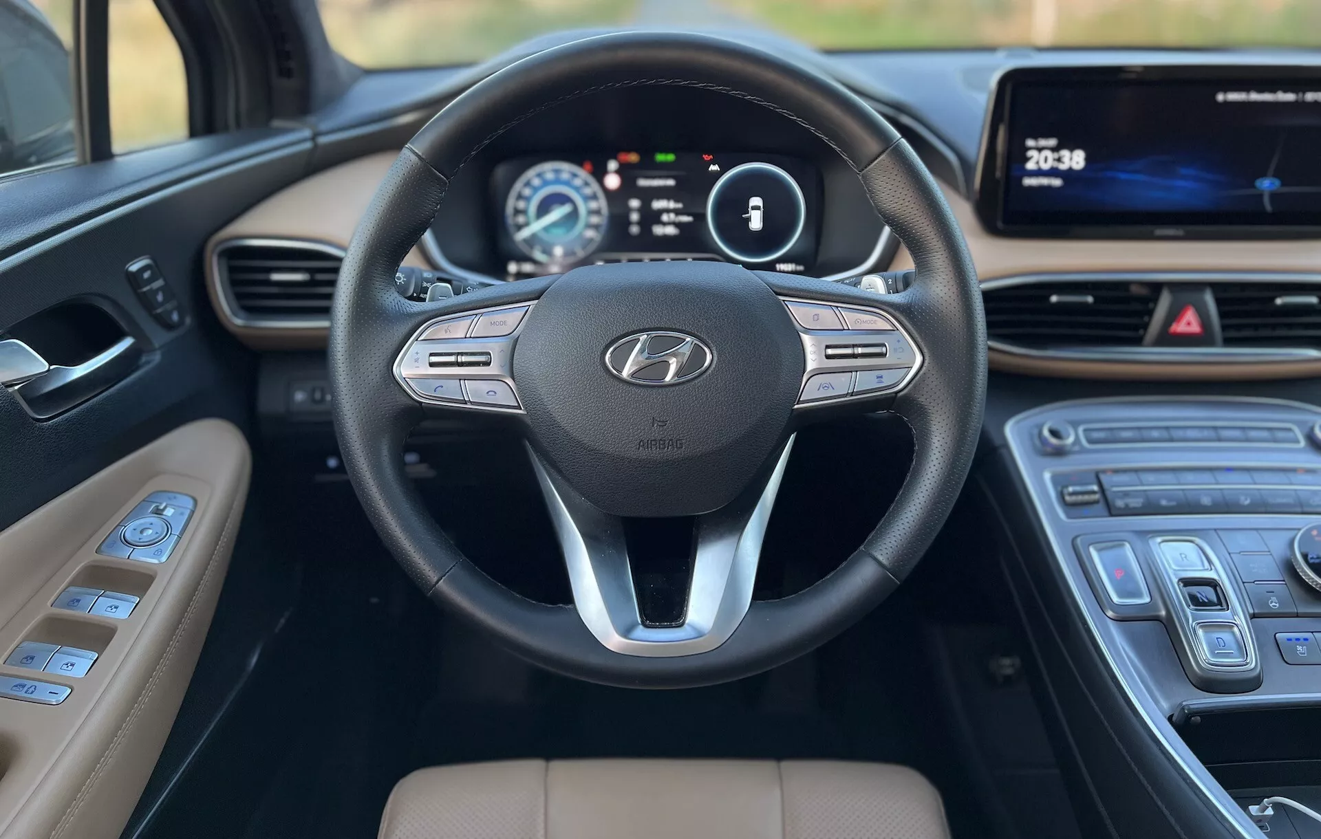 Close-up do ponto de vista dos assentos traseiros de um Hyundai Santa-Fe 2020, focando no painel frontal e volante. O volante possui um design de três raios com o emblema da Hyundai e botões de controle para áudio, comando de voz e controle de cruzeiro. Atrás do volante, um painel de instrumentos digital exibe a velocidade e o RPM em uma tela colorida. A consola central inclui um display touchscreen multimídia e um controle de clima elegante com botões sensíveis ao toque. A alavanca de câmbio tem um design moderno com detalhes prateados, e o estofamento marrom claro contrasta com o painel escuro e o volante, mostrando um interior focado em tecnologia e conforto.