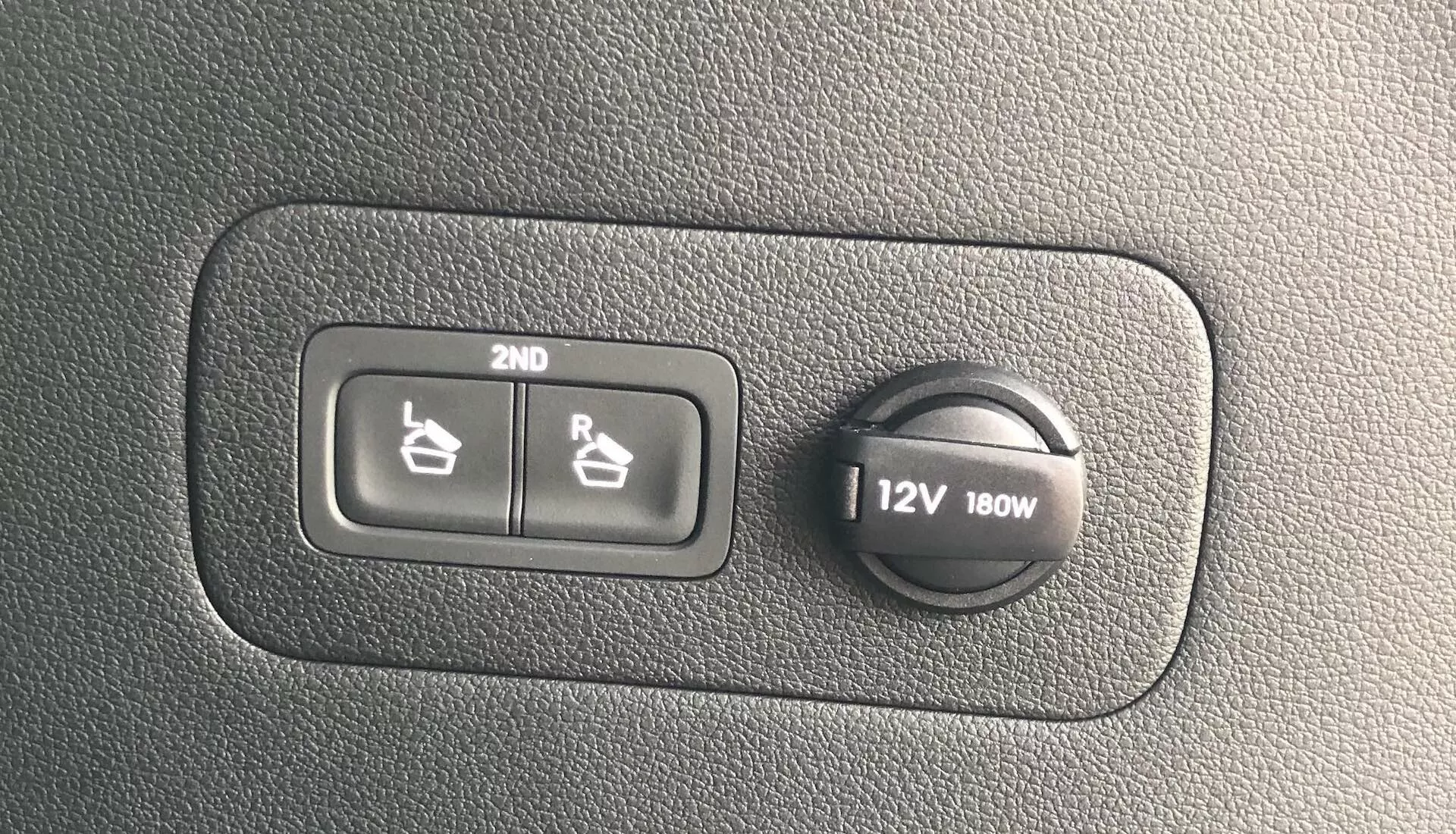 Close-up de uma seção do painel do Hyundai Santa Fe 2020 mostrando os controles do aquecedor de assentos para a segunda fileira e uma tomada de corrente de 12V 180W em uma superfície de plástico texturizado.