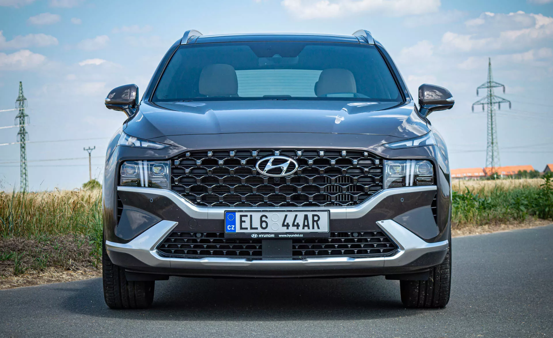 Imagem frontal de um SUV moderno Hyundai Santa Fe 2020 tirada durante o dia. A imagem destaca uma grande grade em cascata com o logotipo central da Hyundai, placa de estilo europeu, elegantes faróis de LED angulares, ampla entrada de ar central e acentos prateados no para-choques inferior. Visíveis estão as linhas esculpidas do capô, para-brisa parcial e espelhos laterais com setas integradas. A imagem enfatiza o tamanho do carro, detalhando seu design moderno e características tecnológicas.