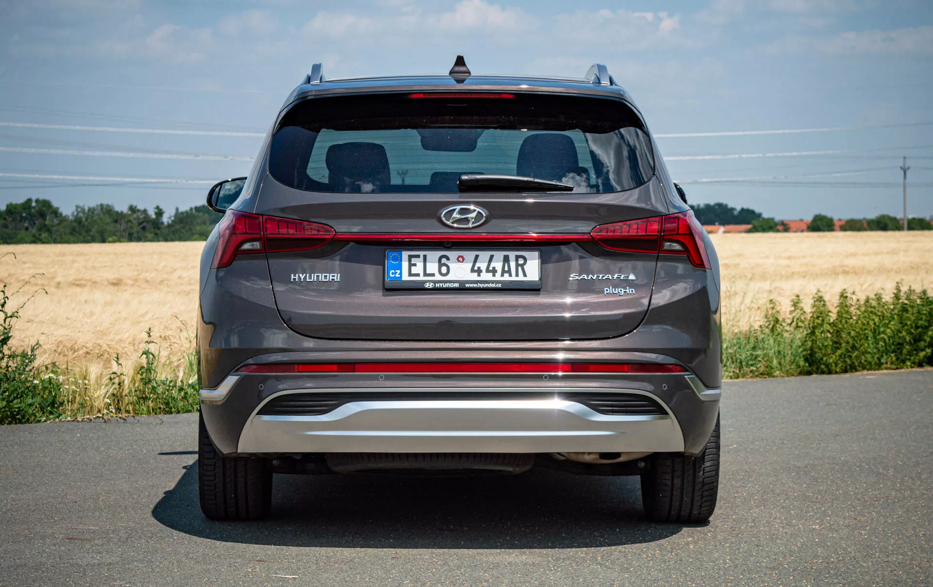 Vista traseira de um moderno SUV Hyundai Santa Fe 2020 prateado-cinza estacionado em uma estrada pavimentada, mostrando o logotipo da Hyundai, o nome do modelo Santa Fe, a placa de estilo europeu, luzes traseiras, para-choques traseiro metálico reflexivo com sensores de estacionamento integrados, pontas de escape duplas, vidro traseiro com spoiler Hyundai e terceira luz de freio. Clima claro com um campo ao fundo.