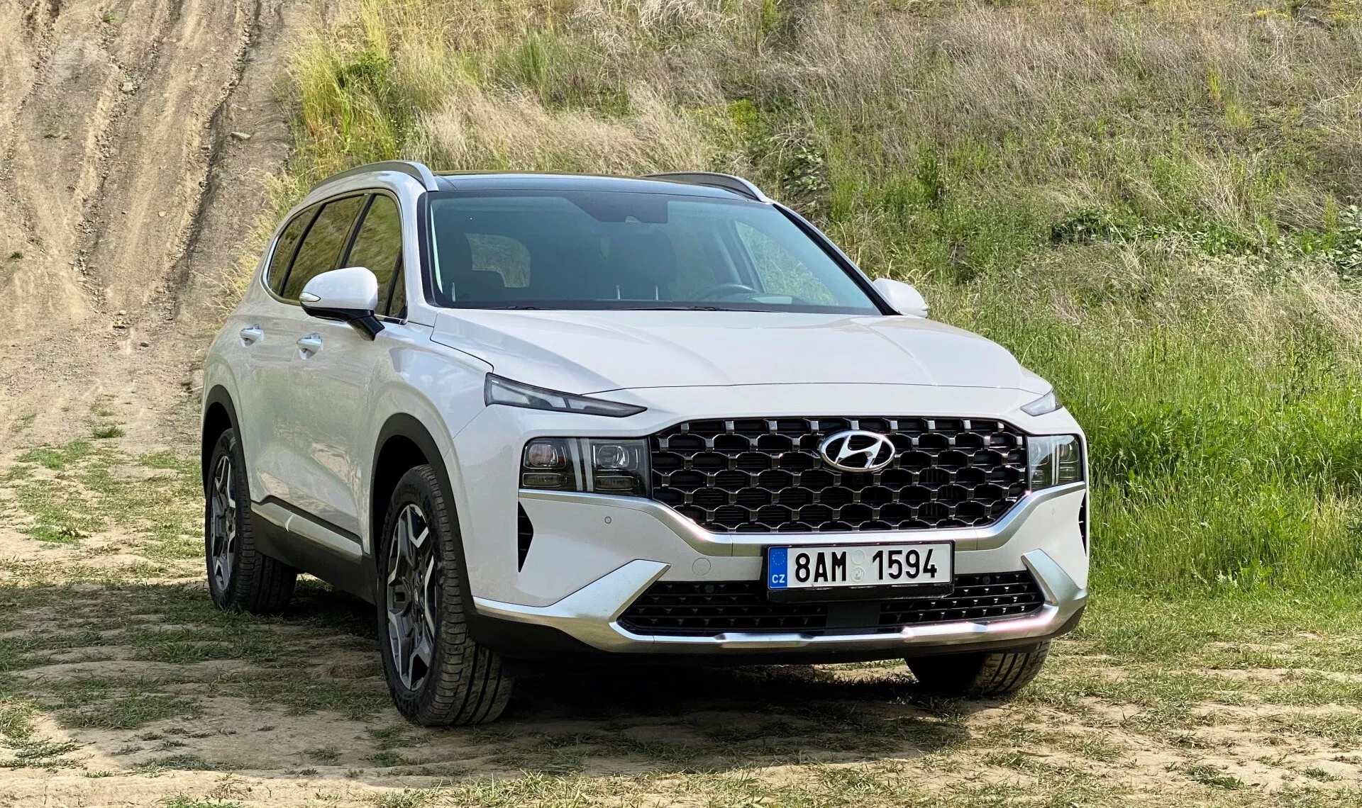 A imagem mostra um SUV Hyundai Santa Fe 2020 branco estacionado em uma superfície não pavimentada com grama curta e um fundo elevado de terra, sugerindo um ambiente de off-road leve. Capturada de uma vista frontal de três quartos, a imagem detalha claramente a fachada frontal do veículo, incluindo uma grade geométrica proeminente que se estende por quase toda a largura, centrada com o logotipo da Hyundai e ladeada por elegantes faróis LED. O para-choque dianteiro inferior abriga iluminação adicional ou sensores. Do perfil lateral até a porta traseira do passageiro, grandes rodas de liga leve preenchem os arcos das rodas, e a carroceria superior apresenta molduras que sugerem robustez. A silhueta do SUV mostra uma linha de teto descendente indicativa de designs esportivos modernos. A cena é naturalmente iluminada, destacando as características do SUV sem sombras fortes.