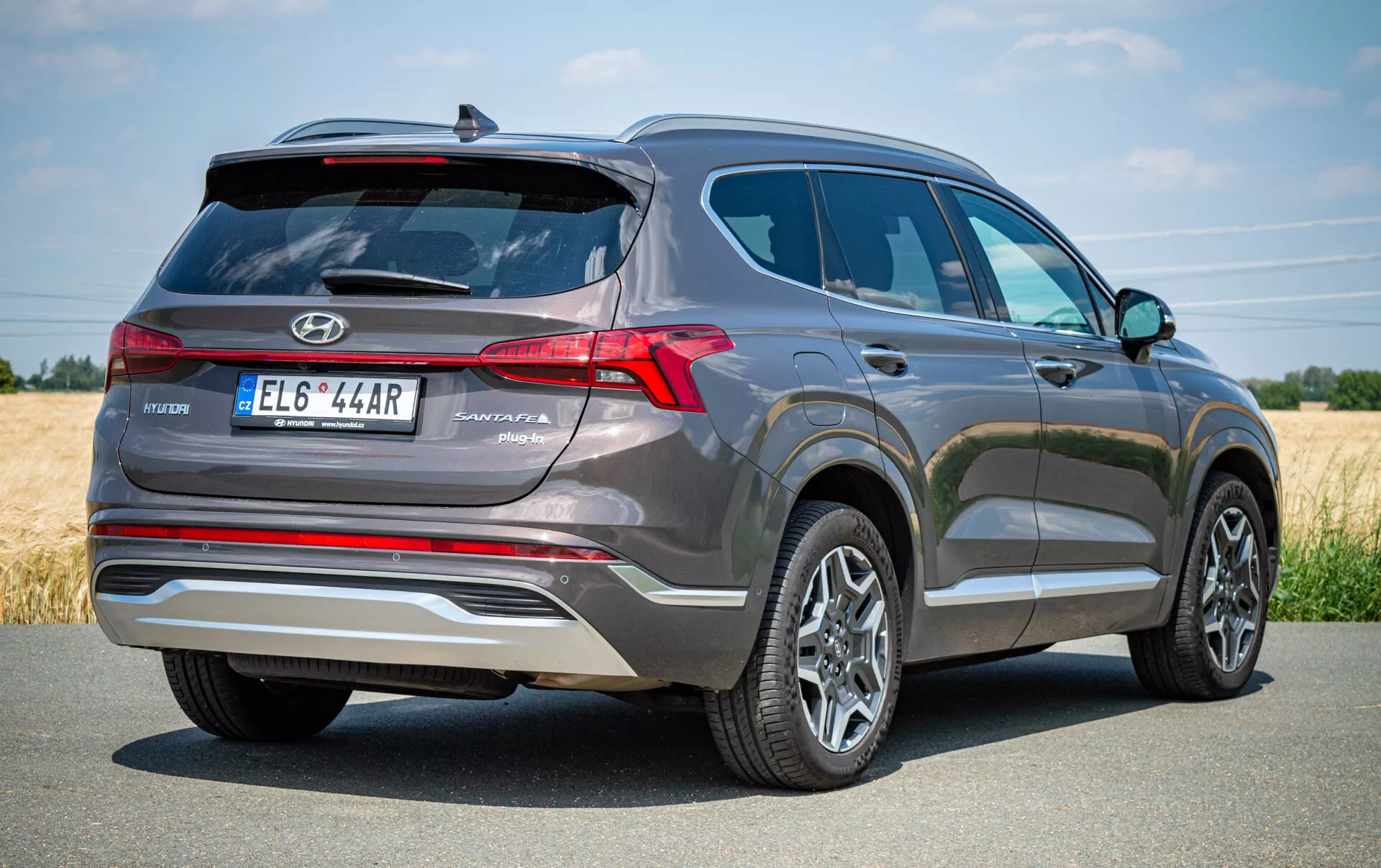 Vista traseira de três quartos de um SUV Hyundai Santa Fe 2020 cinza, destacando o lado traseiro esquerdo. A imagem mostra luzes traseiras LED proeminentes, lavador de para-brisa traseiro, desembaçador e limpador, com emblema da Hyundai e o nome do modelo 'Santa Fe' perto da placa centralizada. Um acabamento prata adiciona estilo, e uma câmera de estacionamento ou sistema de sensores aparece acima da placa. Também são visíveis rodas de liga leve multirraios e pneus em boas condições, indicando um veículo moderno e bem mantido. Ideal para listas automotivas, avaliações ou conteúdo focado em tecnologia.