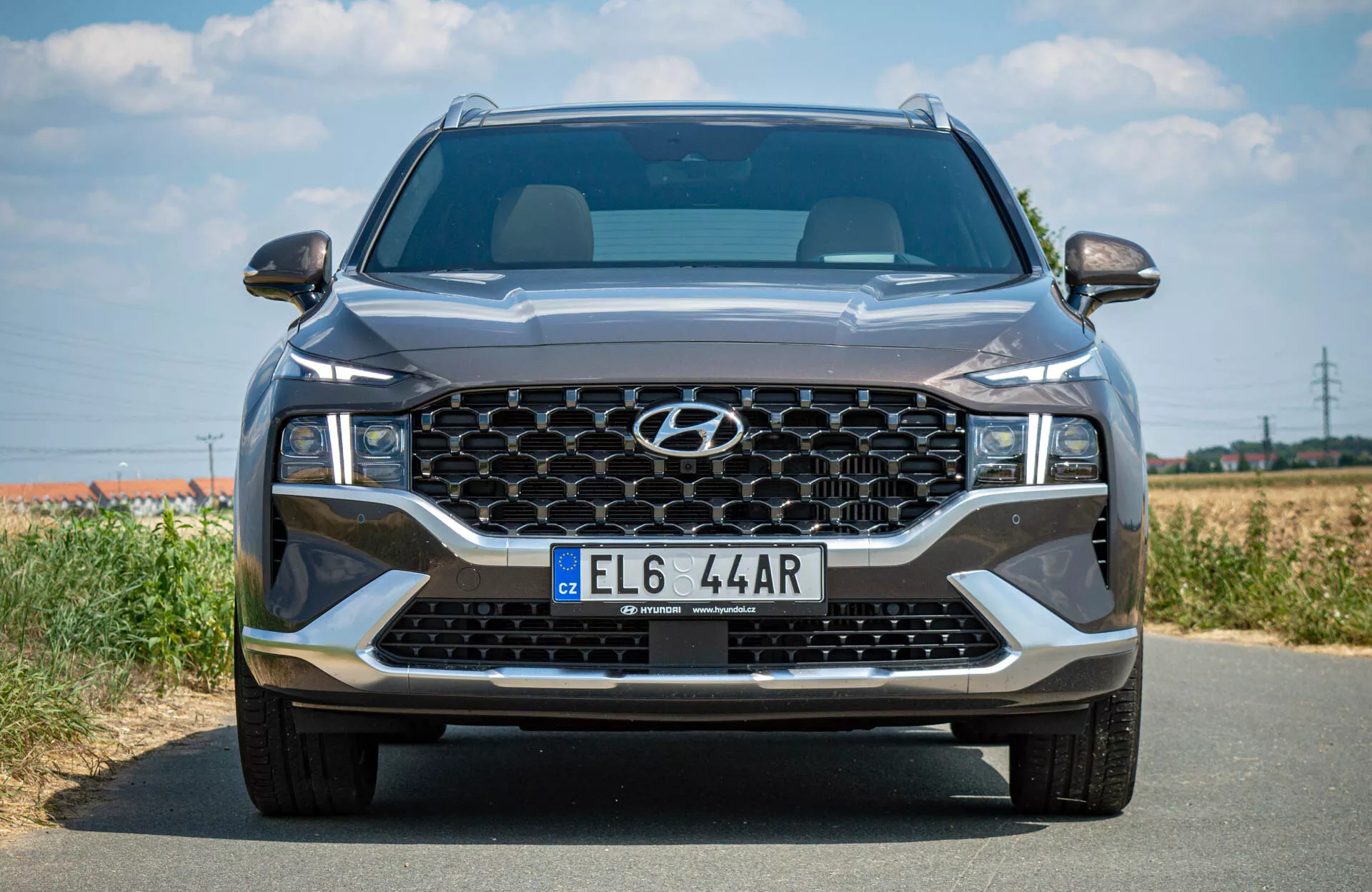 Vista frontal de um SUV Hyundai Santa-Fe 2020 em uma estrada de asfalto, mostrando sua grande grade hexagonal com padrão de favo de mel, faróis LED elegantes, emblema centralizado na parte superior da grade, luzes de funcionamento diurno em LED abaixo dos faróis principais e placa de licença de estilo europeu na parte inferior da grade. O fundo é uma paisagem rural ou semi-rural desfocada com um céu claro, indicando boas condições meteorológicas.