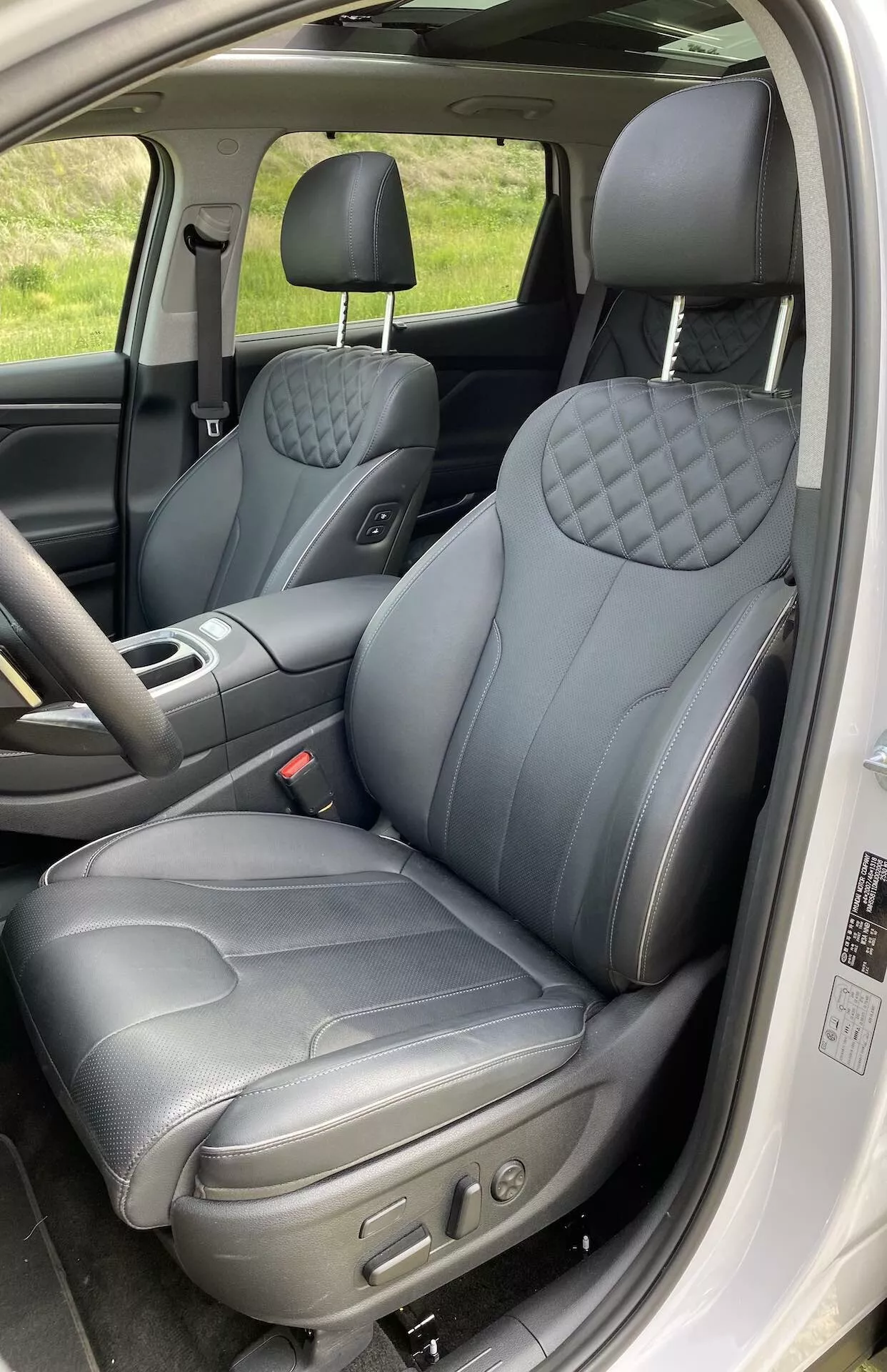 Interior do Hyundai Santa Fe 2020 mostrando o assento do motorista a partir de uma porta aberta. A foto vertical de perto captura os bancos dianteiros, focando no banco do motorista que apresenta estofamento de couro escuro com padrão acolchoado em forma de diamante no encosto e no assento, sugerindo uma variante premium. Controles eletrônicos visíveis indicam posições ajustáveis do assento. O apoio de cabeça parece ajustável, e o painel da porta possui controles de janelas e possivelmente de espelhos. O interior é detalhado com costuras contrastantes, realçando a sensação de luxo.