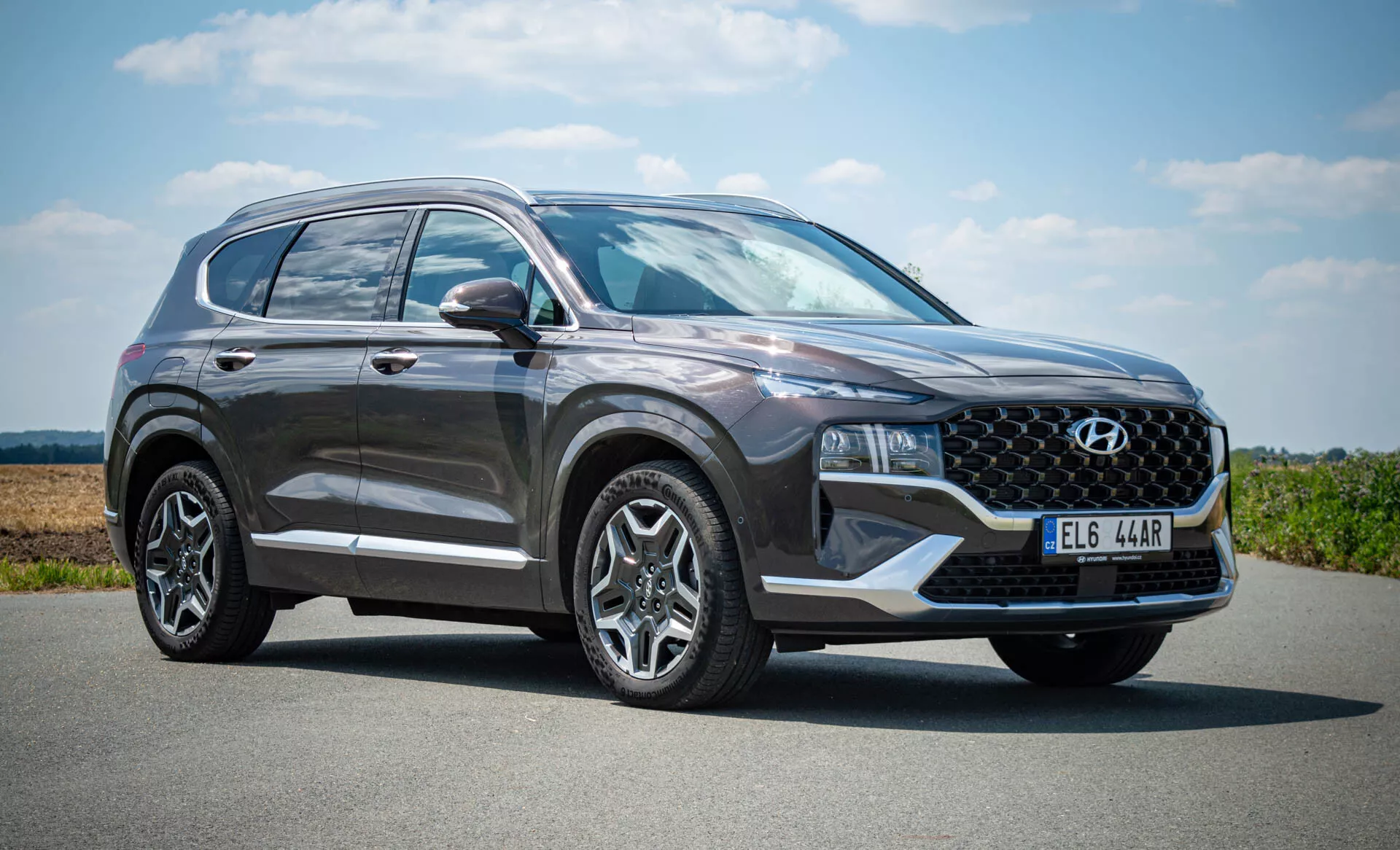 Hyundai Santa Fe 2020 SUV estacionado em uma superfície plana sob um céu claro com vegetação ao longe. A visão do quarto dianteiro esquerdo destaca sua grade proeminente, faróis de LED elegantes, para-choque dianteiro, porta do motorista, espelhos laterais com indicadores de direção integrados, rodas de liga leve e uma parte da janela traseira e lanterna traseira. O acabamento metálico da cor escura do carro é realçado pela luz natural, sugerindo um dia ensolarado.