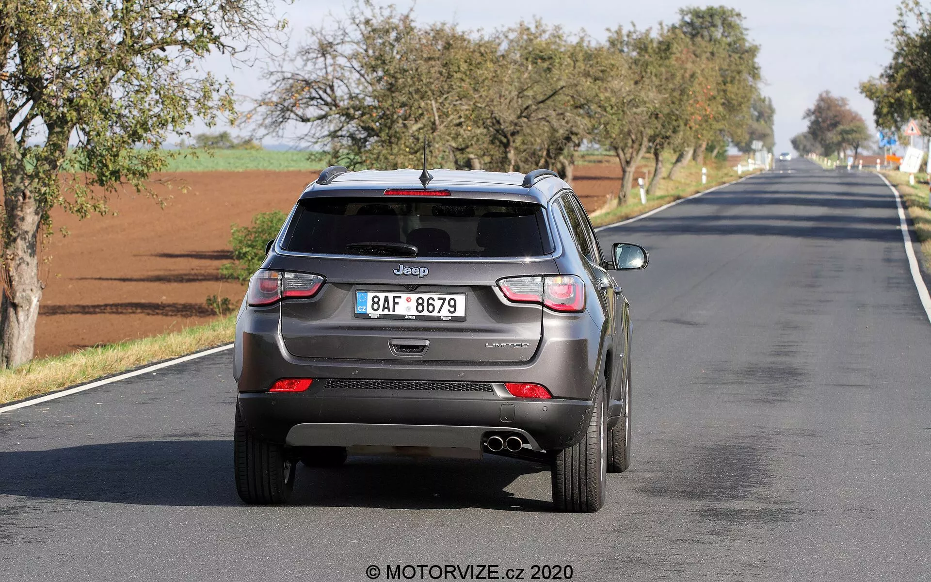 Vista traseira de um Jeep Compass 2017 a dirigir-se numa estrada de duas faixas, exibindo elementos de design moderno, como antena estilo barbatana de tubarão, limpador de para-brisa traseiro, luzes traseiras em LED, sensores de estacionamento integrados, tubos de escape duplos e uma placa de matrícula de estilo europeu com o logotipo da Jeep claramente visível. O veículo parece estar em movimento, capturando uma operação em tempo real com tecnologias de assistência ao condutor potenciais.
