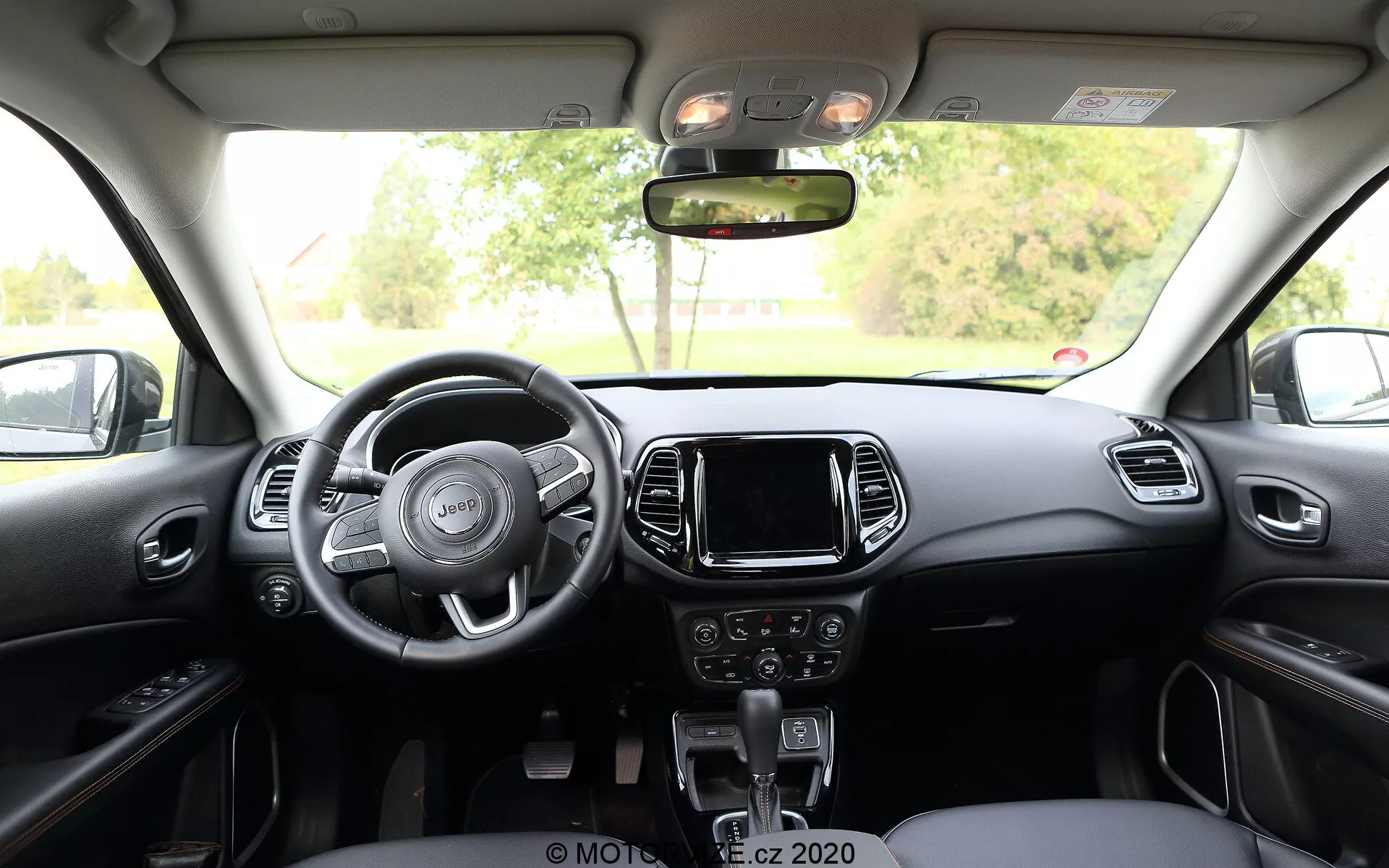 Vista interior do Jeep Compass 2017 a partir do banco traseiro, exibindo um volante de três raios com controles integrados, console central com sistema de infoentretenimento touchscreen, controles manuais para áudio e clima abaixo dele, e uma alavanca de câmbio automático. O painel e os assentos dianteiros apresentam um esquema de duas cores com detalhes metálicos ou plásticos, enquanto nas partes superiores há controle de luz da cabine e teto solar.