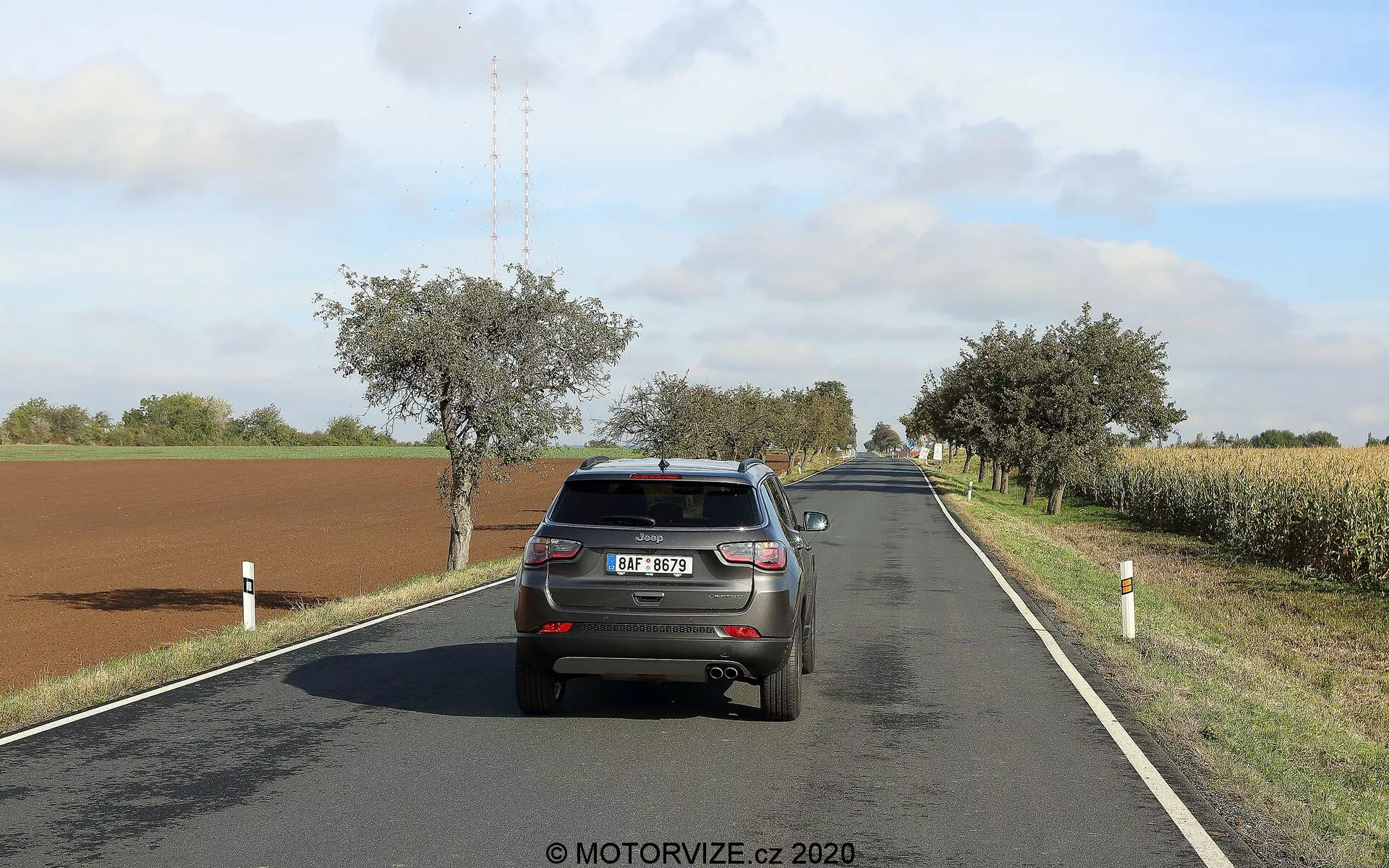A imagem mostra a vista traseira de um Jeep Compass 2017 dirigindo em uma estrada rural de duas faixas em um dia ensolarado com nuvens dispersas. O veículo, posicionado no lado direito da pista, exibe seu portão traseiro, para-choque, luzes traseiras, para-brisa traseiro, placa de licença, parte do teto com a antena de barbatana de tubarão e a parte superior dos pneus traseiros. O carro ocupa uma parte significativa do quadro, permitindo um detalhamento claro de suas características. A estrada curva-se suavemente para a direita à distância, cercada por campos, árvores e postes à beira da estrada. As luzes de freio do carro não estão acesas, sugerindo cruzeiro normal.