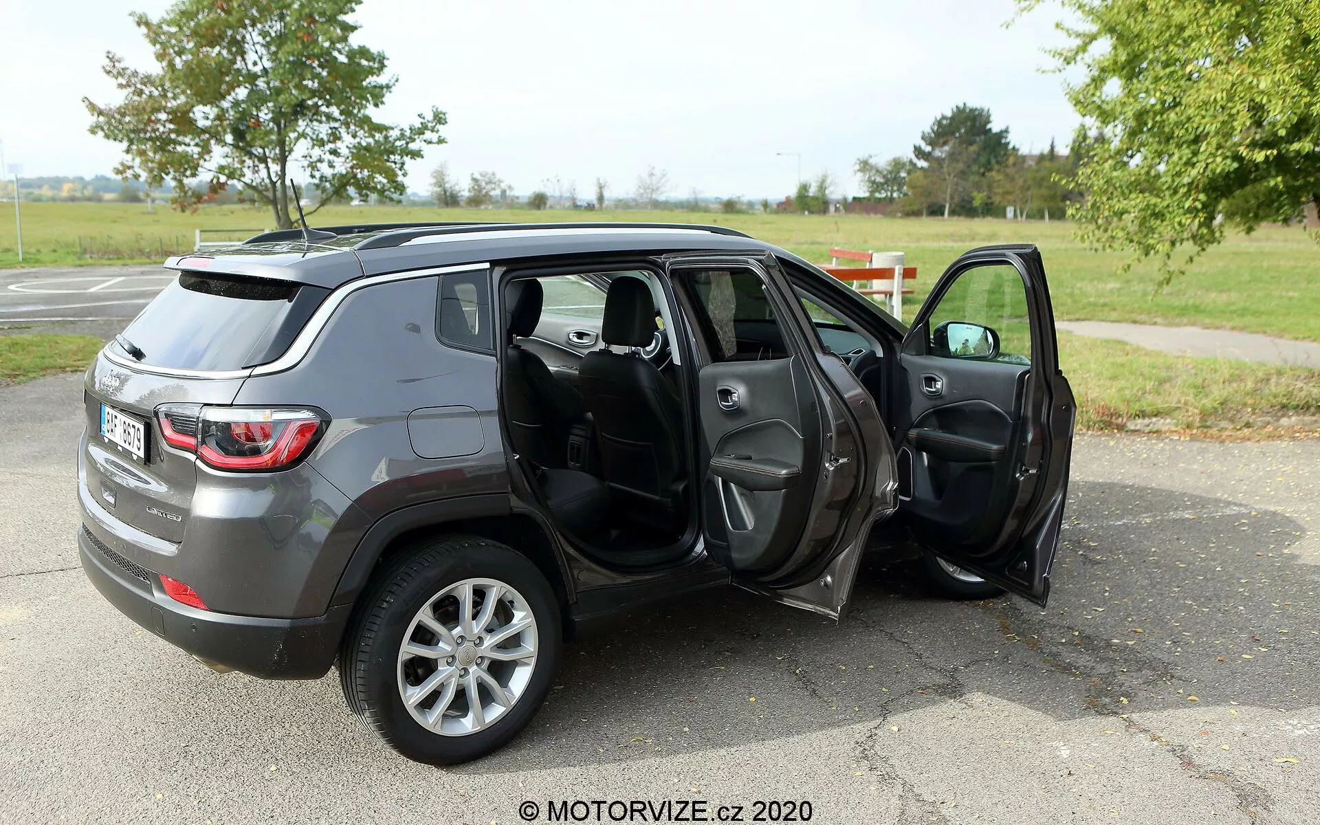 Imagem de um SUV Jeep Compass 2017 de tamanho médio estacionado em uma superfície de asfalto em um ambiente semelhante a um parque com grama e árvores ao fundo. A foto é tirada de um ângulo traseiro de três quartos, mostrando o perfil e a parte traseira do veículo. Ambas as portas do lado do passageiro estão abertas, revelando o interior de tecido ou couro escuro, incluindo os bancos dianteiros e traseiros, com possíveis recursos como janelas e espelhos elétricos. O SUV tem acabamento metálico cinza e está equipado com rodas de liga leve de cinco raios, o que pode indicar um nível de acabamento mais alto ou uma atualização opcional das rodas. A marca d'água 'MOTORVIZE.cz 2020' sugere que a imagem é de uma revisão de carro ou site automotivo.