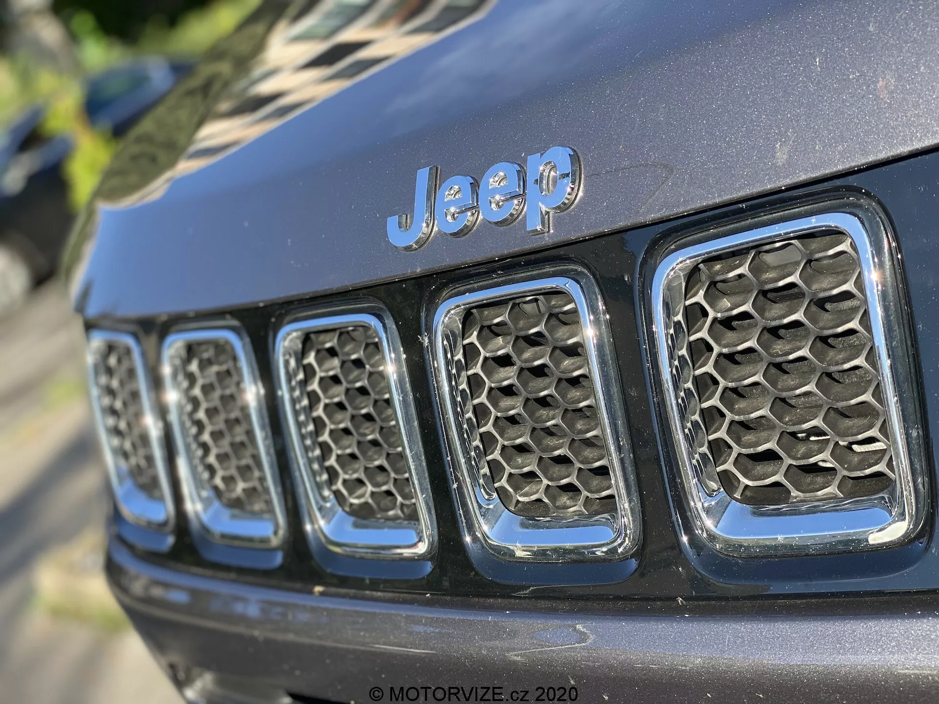 Vista de close-up da grade frontal de um Jeep, mostrando o emblema 'Jeep' proeminente centralizado acima das sete ranhuras verticais, um elemento de design característico. A fotografia é tirada em um ângulo oblíquo, focando de perto no lado esquerdo da grade e de uma perspectiva ligeiramente baixa. As ranhuras da grade têm um padrão de favo de mel, sugerindo melhor fluxo de ar e proteção para o radiador e componentes do motor. O acabamento de pintura preta brilhante indica um veículo relativamente novo ou bem mantido. A baixa profundidade de campo mantém o emblema 'Jeep' e algumas ranhuras em foco nítido, enquanto o resto se desfoca, destacando os elementos de design e marca sob iluminação natural ao ar livre com sombras suaves.
