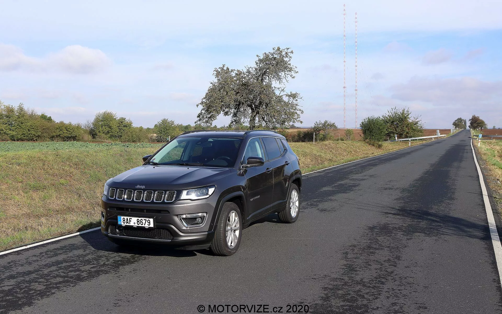 Imagem de um Jeep Compass 2017 fotografado de um ângulo frontal de três quartos. O veículo, posicionado à direita de uma estrada de duas faixas, exibe seu perfil frontal e lateral. Os recursos principais incluem uma grade moderna, faróis proeminentes com luzes diurnas de LED, rodas de liga leve, sinalizadores de direção integrados nos retrovisores, uma antena no teto e faróis de neblina no para-choque dianteiro. O ambiente parece rural, com céu claro e vegetação ao longo da estrada.