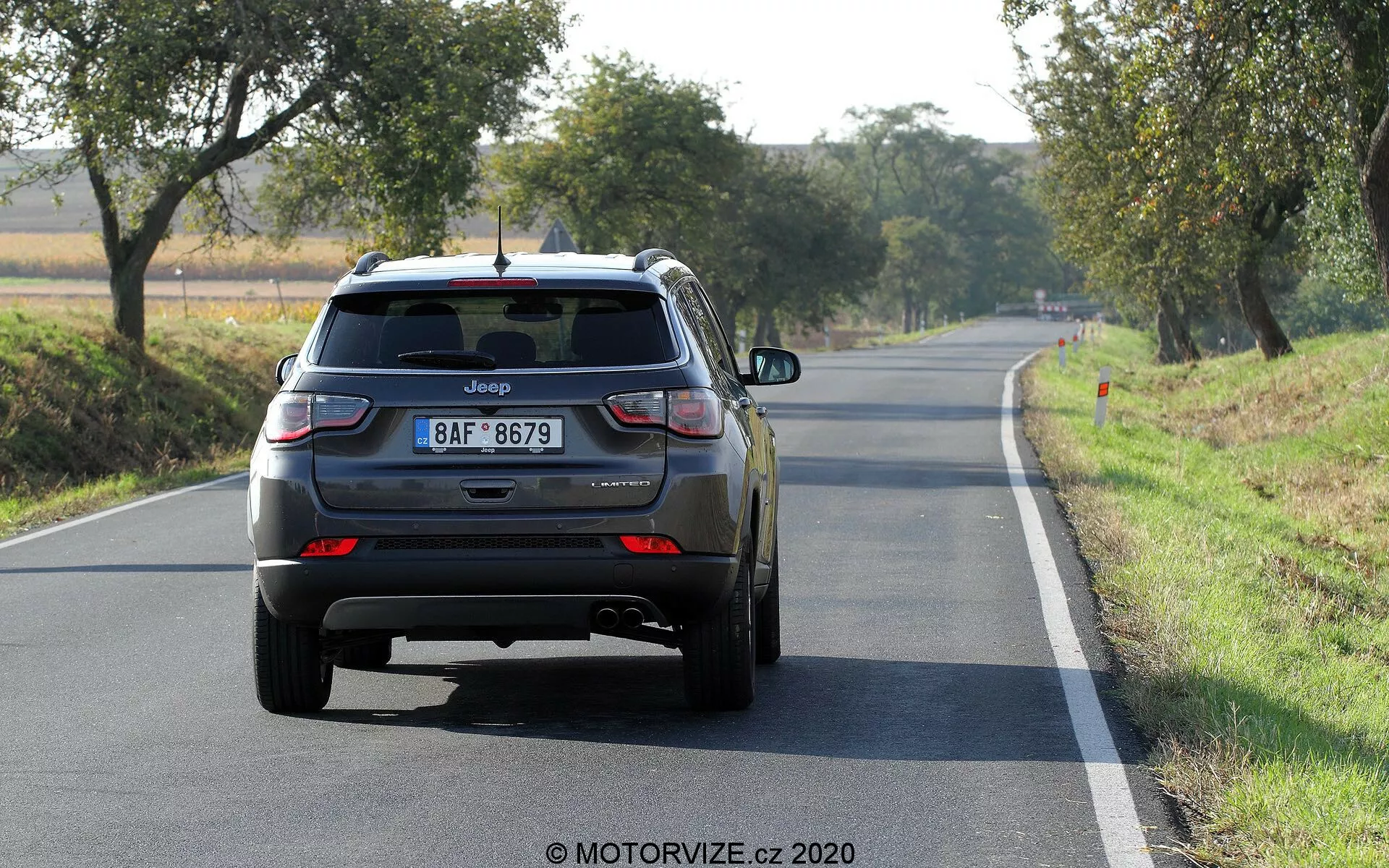 Vista traseira de um Jeep Compass SUV 2017 em uma estrada de duas pistas à luz do dia, com logotipo visível, nome do modelo, placa de estilo europeu, luzes traseiras de LED, porta traseira fechada com limpador de para-brisa traseiro, terceira luz de freio no spoiler traseiro, para-choque traseiro com refletores, tubos de escape duplos no lado esquerdo, rodas traseiras de liga, emblemas adicionais possivelmente indicando o nível de acabamento ou transmissão.