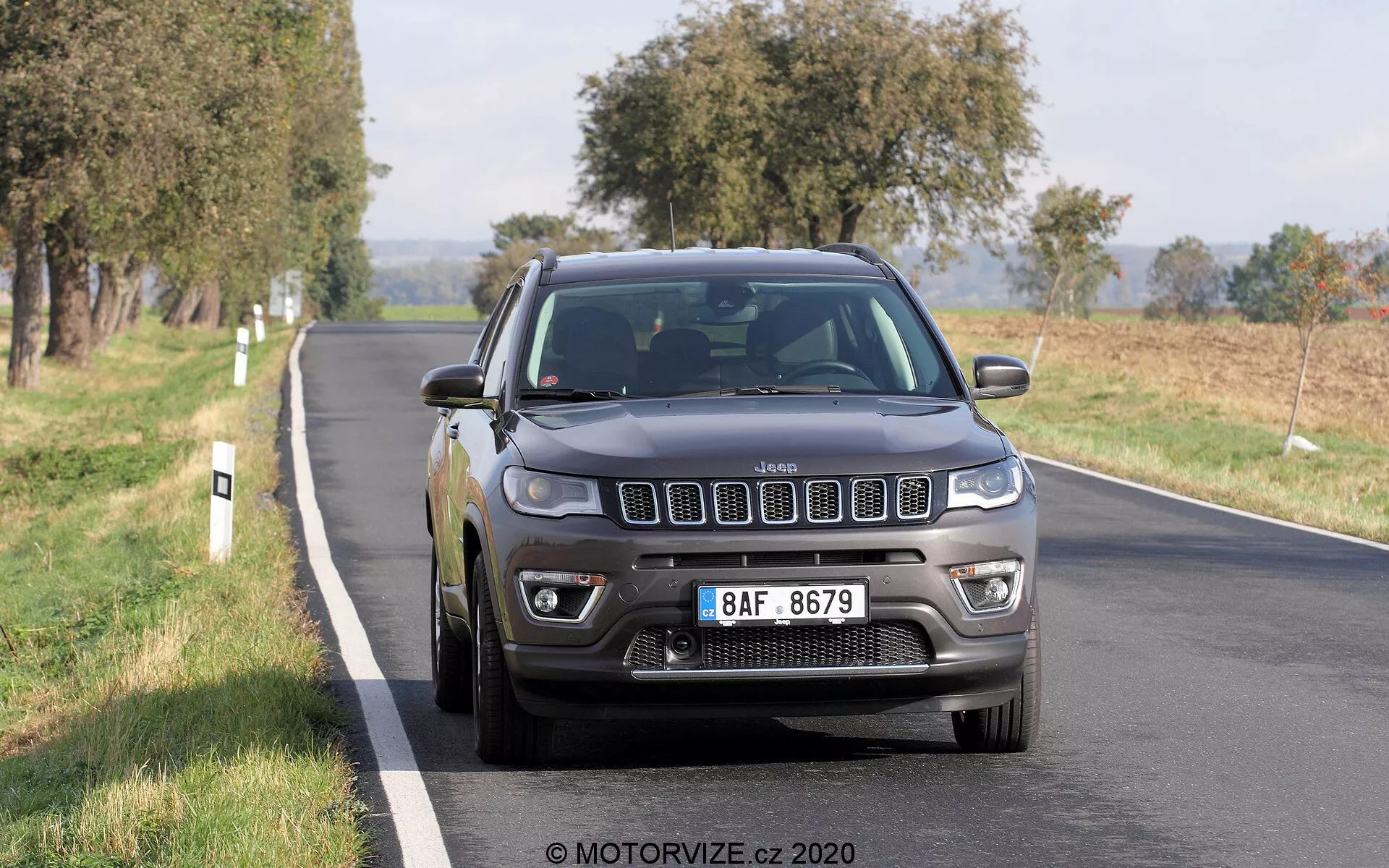 Imagem de um Jeep Compass 2017 dirigindo em uma estrada de duas faixas, fotografado em um ângulo frontal de três quartos. A icônica grade de sete fendas do veículo, os faróis detalhados com luzes diurnas LED, os faróis de neblina e as rodas de liga leve multi-raios são claramente visíveis. A cor cinza escuro ou preto do Jeep dá-lhe uma aparência sólida e robusta, com luz natural do dia destacando suas características. O veículo parece estar em movimento, com um leve desfoque nas rodas. Árvores e campos abertos no fundo sugerem um ambiente rural ou semi-rural. O carro ocupa a metade central e inferior da moldura, ligeiramente à direita, capturando sua grade dianteira, faróis, para-choque, capô, para-lama lateral do motorista, roda e porta dianteira.