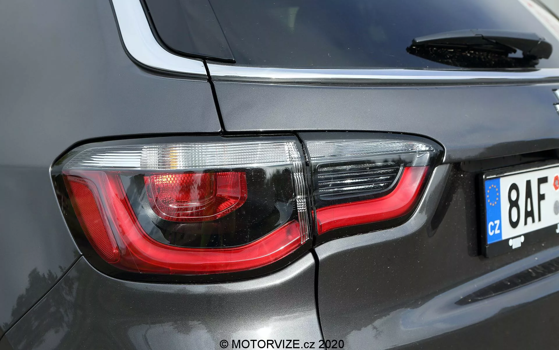 Imagem aproximada da seção traseira direita de um Jeep Compass 2017 preto ou cinza escuro, destacando um design moderno da luz traseira com lentes claras e vermelhas, tecnologia LED e material refletivo de cromo para melhor visibilidade. A imagem mostra parte do pára-brisa traseiro, a antena e uma placa de licença de estilo europeu, capturada de um ângulo diagonal e fora do centro, destacando as curvas e o estilo contemporâneo do SUV.