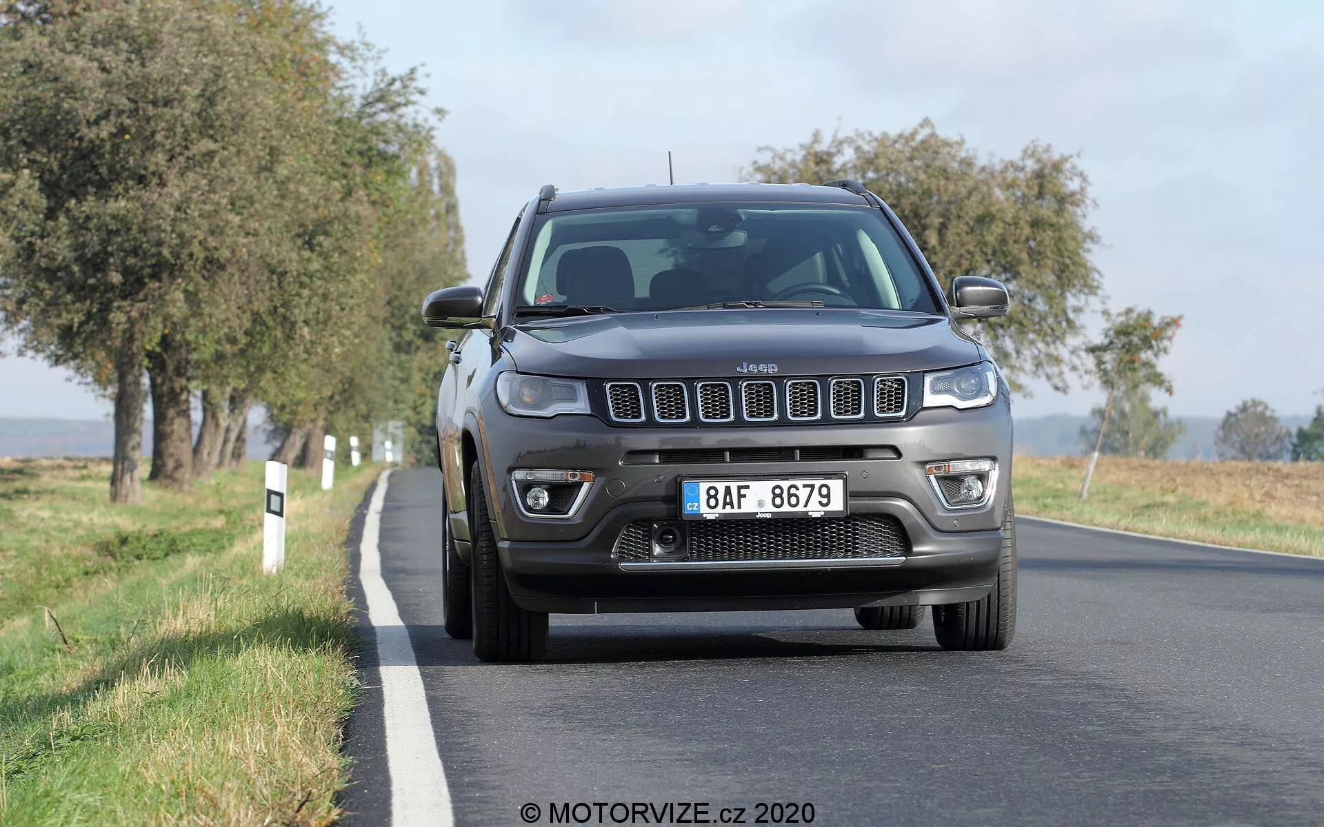 Vista frontal de um Jeep Compass 2017 dirigindo em uma estrada rural com árvores alinhadas em ambos os lados. O veículo apresenta a distintiva grade de sete fendas e faróis quádruplos halógenos automáticos com função DRL. O parachoque dianteiro inclui luzes de neblina e uma placa de licença de estilo europeu está montada na frente. A imagem captura o veículo em luz diurna clara, destacando detalhes da grade, faróis, capô e para-lamas.