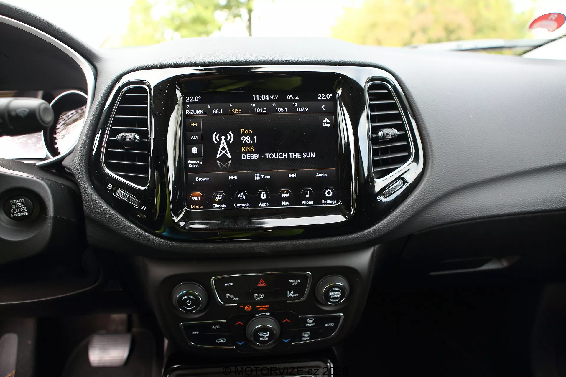 O interior de um Jeep Compass 2017, destacando a área central do painel de instrumentos com uma grande tela sensível ao toque de infotainment central que exibe funcionalidades de conectividade como rádio, mídia e navegação por satélite. Abaixo da tela sensível ao toque encontram-se os controles do sistema de climatização, incluindo botões e botões com detalhes cromados e um display LCD. Os controles montados no volante parcialmente visível incluem opções para controle de cruzeiro e ajuste do volume do áudio. Também são mostrados detalhes adicionais de design, como saídas de ar verticais, controles de aquecimento dos assentos e uma porta USB, destacando a tecnologia interior e o design estético do veículo.
