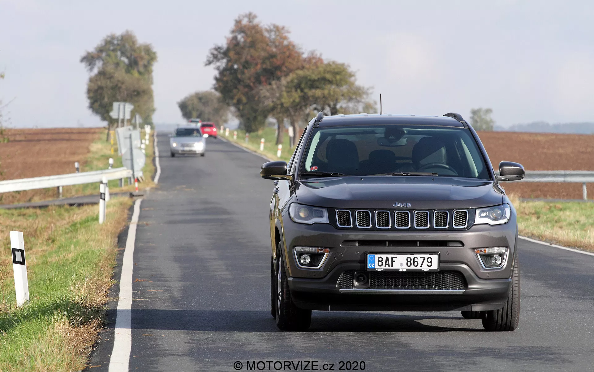Foto de ângulo baixo de um Jeep Compass 2017 dirigindo em direção à câmera em uma estrada pavimentada de duas pistas. A vista frontal mostra a grade, os faróis e o para-choque em detalhe com características modernas visíveis como luzes LED ou HID e sensores de estacionamento frontais. O carro está em movimento, com um fundo rural desfocado apresentando postes brancos na beira da estrada e campos abertos sob um céu claro.