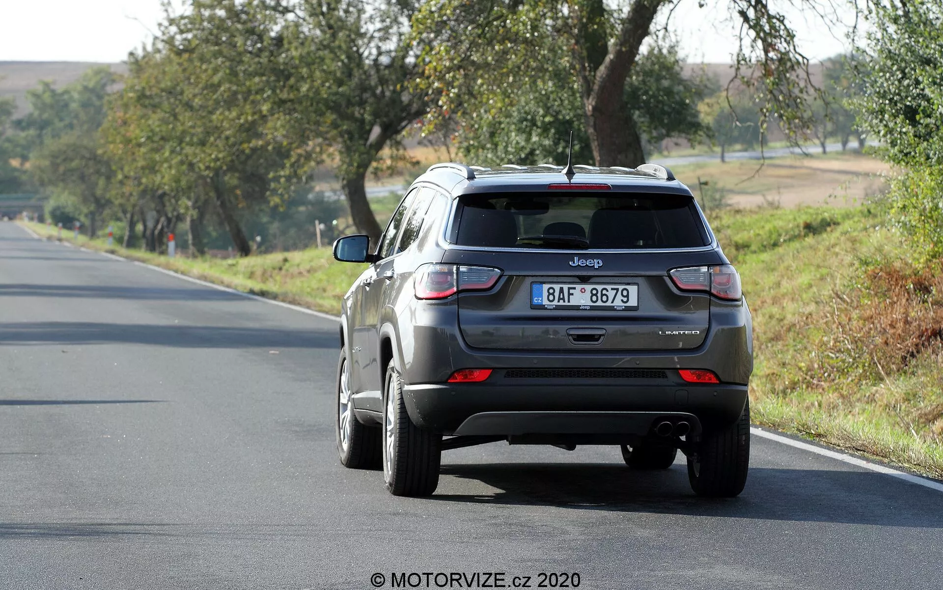 Vista traseira de um SUV Jeep Compass 2017 cinza metálico dirigindo por uma estrada de duas faixas, posicionado ligeiramente à direita do quadro. O veículo, na faixa da direita, tem características visíveis como a porta traseira com o emblema Jeep e a placa de matrícula, o para-brisa traseiro com um pequeno spoiler, luzes traseiras com seções vermelhas e brancas, para-choque traseiro com sensores e uma antena barbatana de tubarão no telhado. As rodas de liga são parcialmente visíveis. A cena sugere um dia ensolarado com árvores e grama ao redor, indicando um ambiente rural ou semi-rural.