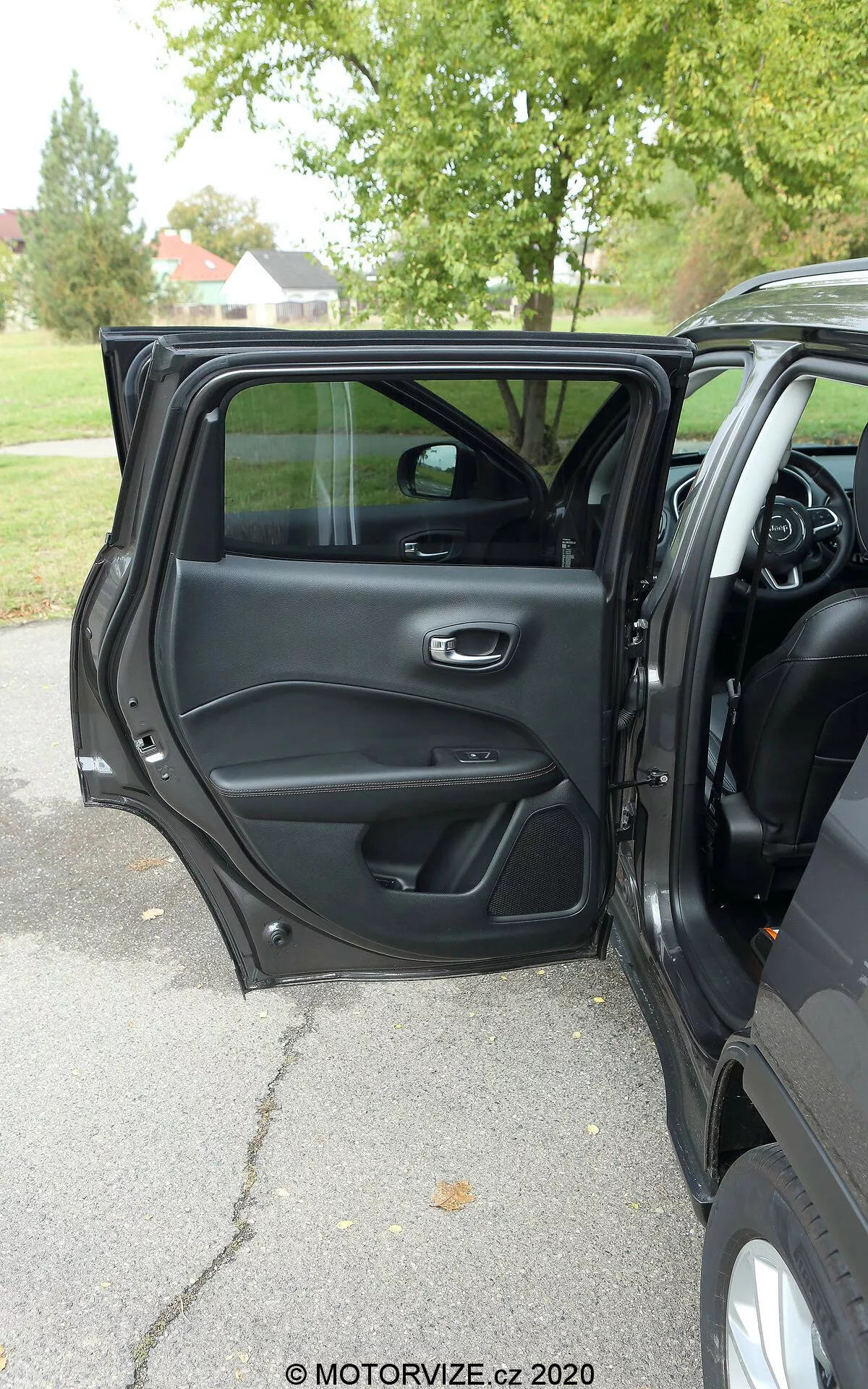 Vista do interior de um Jeep Compass 2017 a partir do exterior, mostrando a porta do lado do motorista aberta com uma maçaneta metálica e ergonômica, compartimentos de armazenamento, grade de alto-falante e painel de controle para operações de janela e travamento de portas. Parte do painel de instrumentos, console central e volante multifuncional são visíveis, sugerindo um interior moderno e rico em recursos. Luz natural, clima nublado, árvores ao fundo sugerindo um ambiente ao ar livre em um parque ou rua arborizada.
